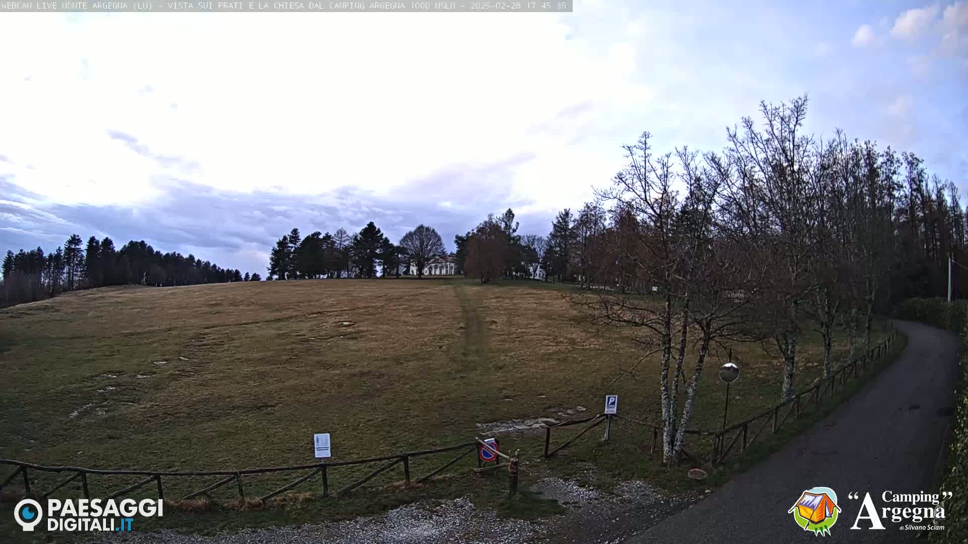 A mostly cloudy sky overlooks a large, flat, brown field bordered by leafless trees and a paved road leading to a wooden gate.