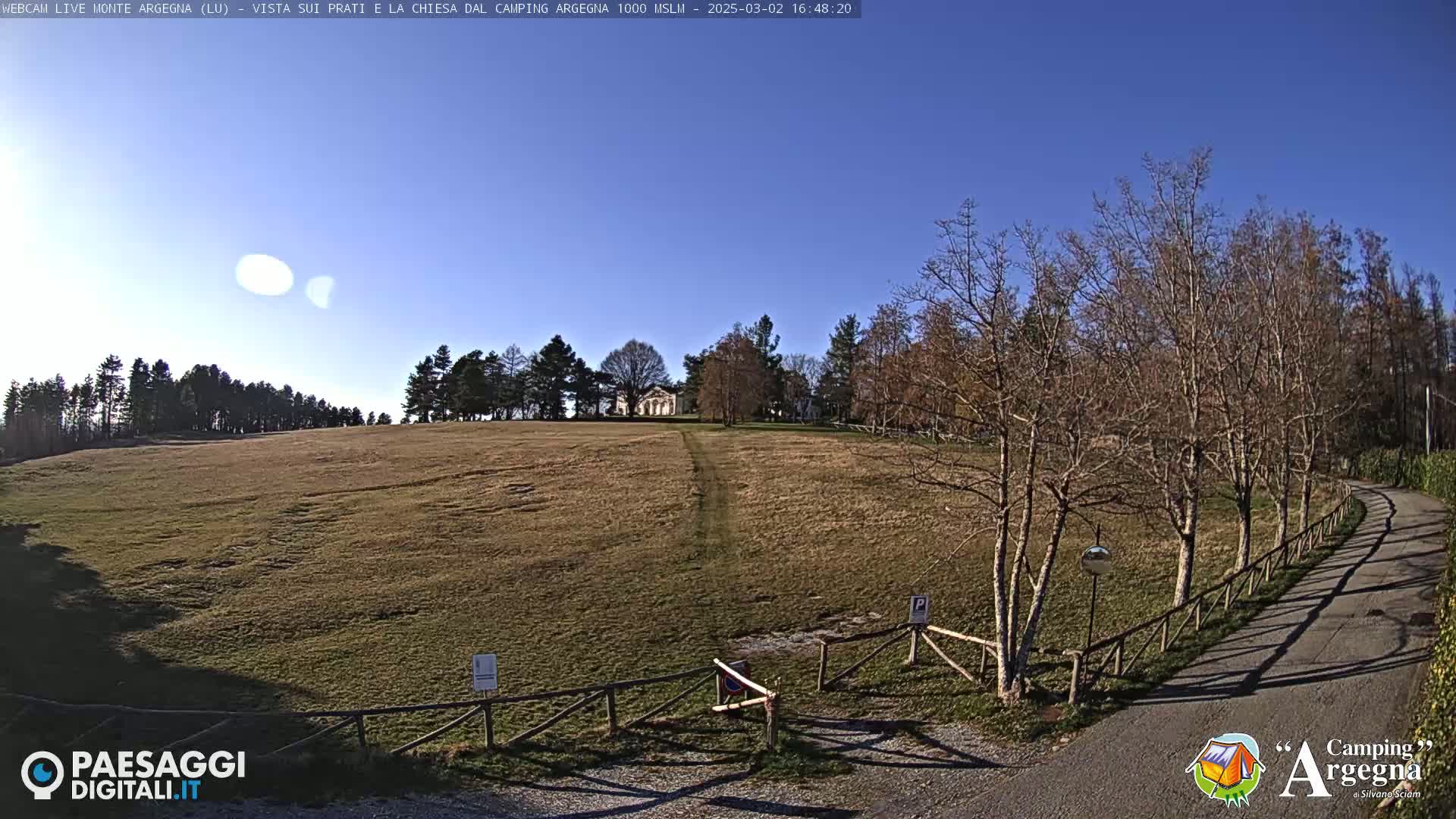 A wide, grassy field slopes gently upward towards a building, framed by leafless trees under a clear blue sky.