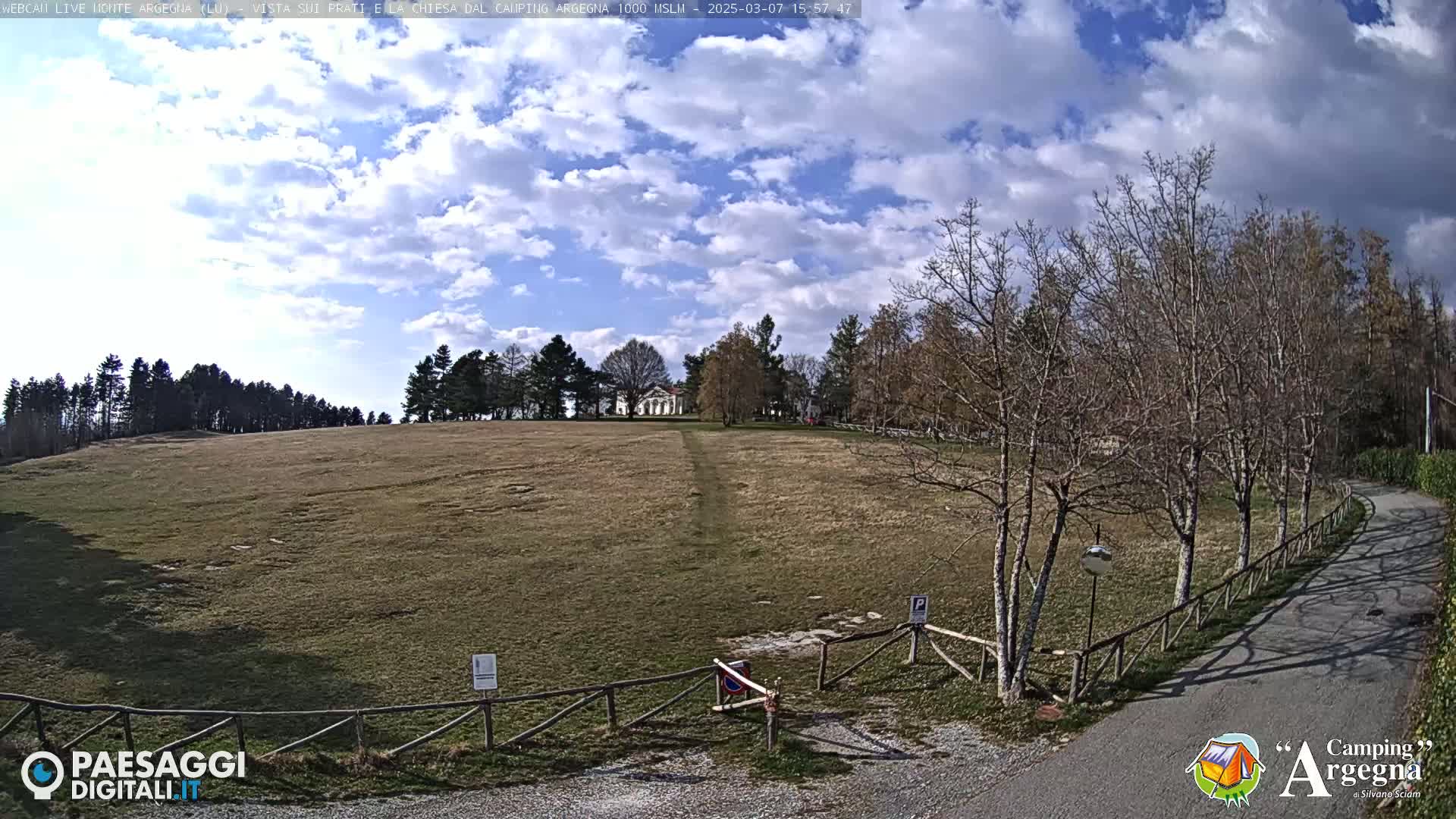 A wide, grassy field stretches towards a distant building, under a partly cloudy blue sky; bare trees line a curving road leading to a wooden gate.