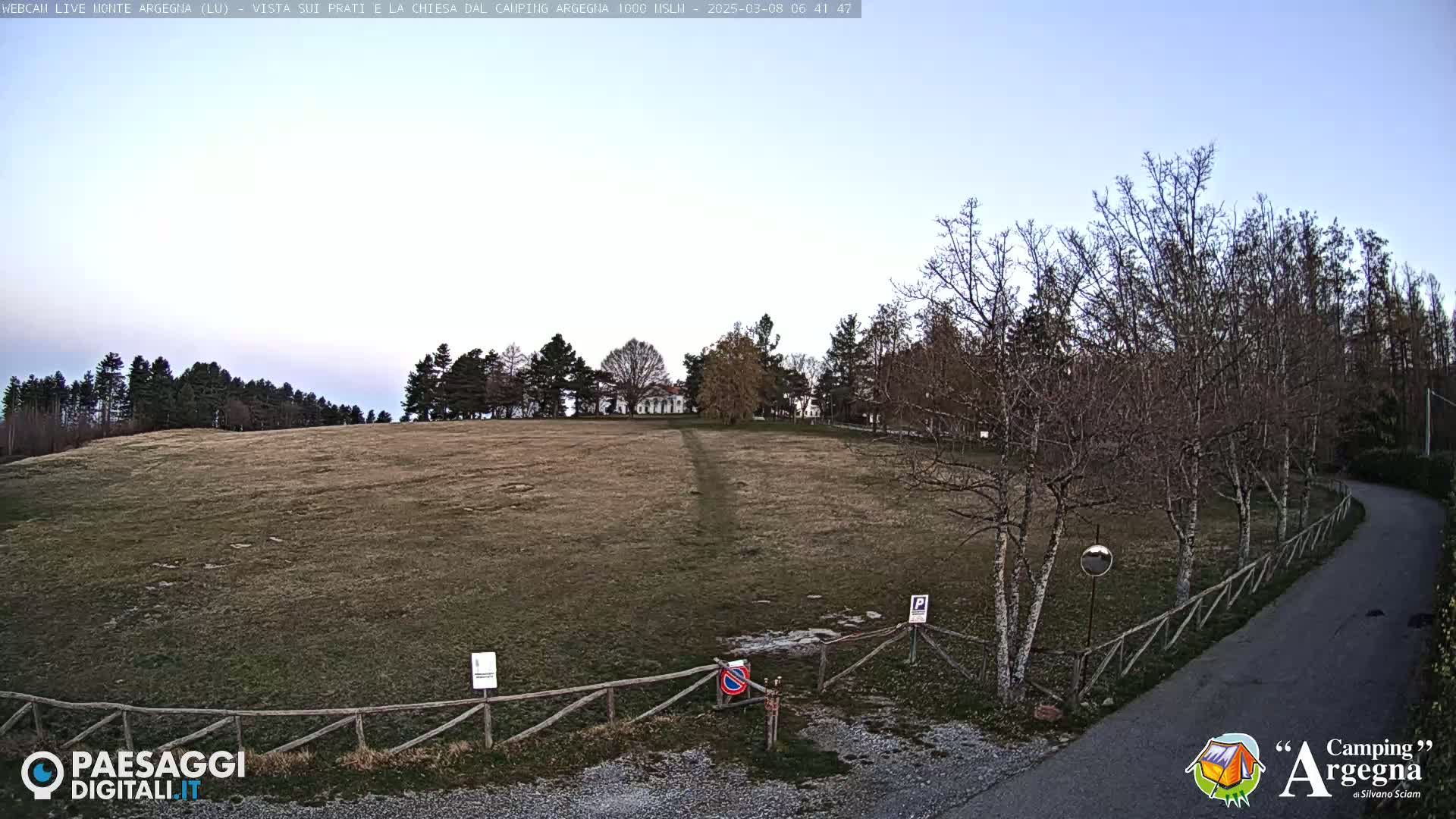 A large, grassy field with leafless trees and a paved road curves gently to the right, under a clear, light-blue sky.