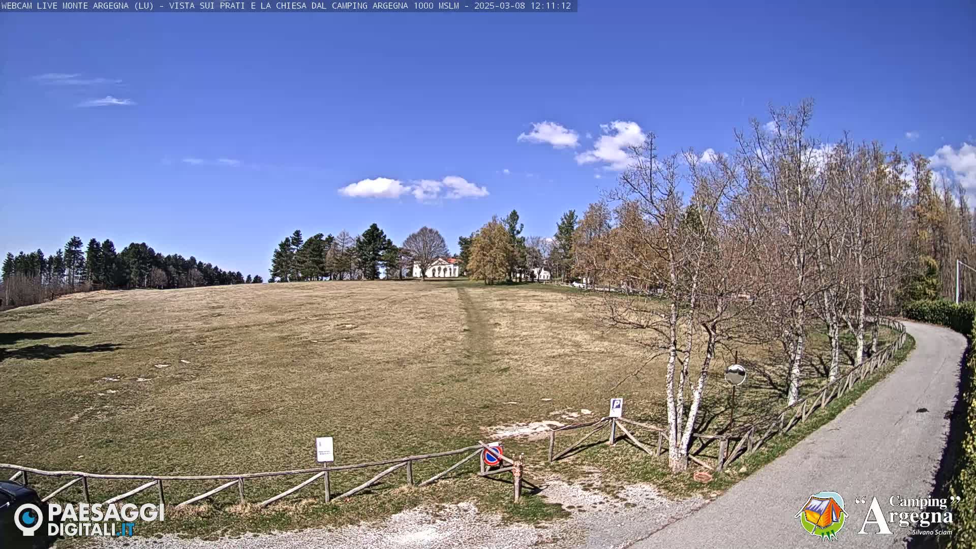 A mostly sunny day reveals a grassy field, a small white building in the distance, and a paved road curving through bare trees beside a wooden fence.
