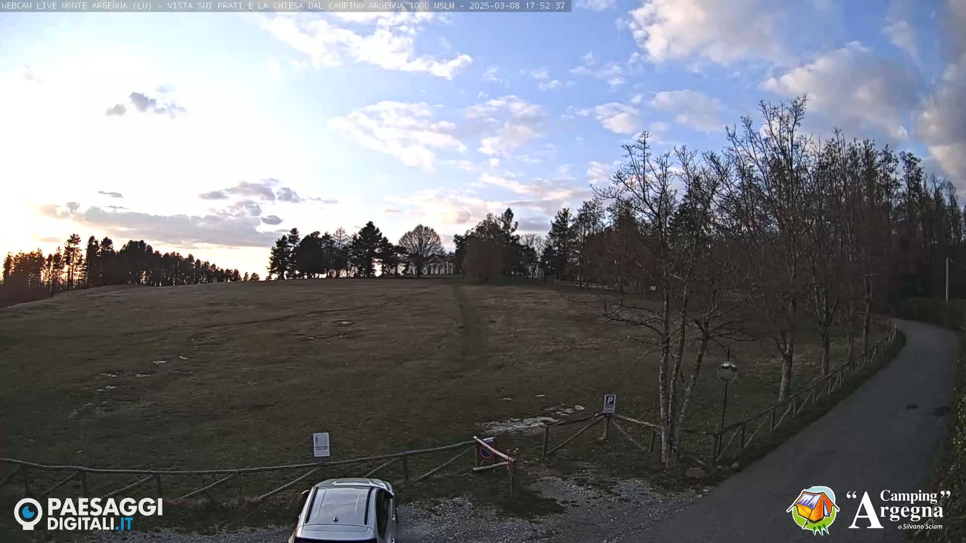 A mostly-clear sky over a large, flat, grassy field bordered by leafless trees and a paved road, with a car parked near a wooden fence.
