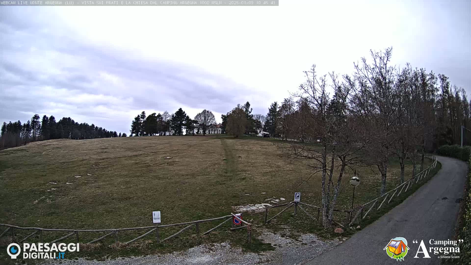 A large, grassy field stretches out under an overcast sky, with bare trees and a paved road leading toward a building in the distance.