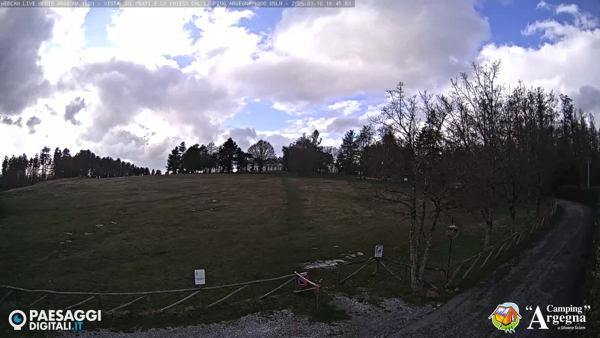 A mostly cloudy sky over a grassy field, with bare trees and a dirt road leading to a wooden fence.