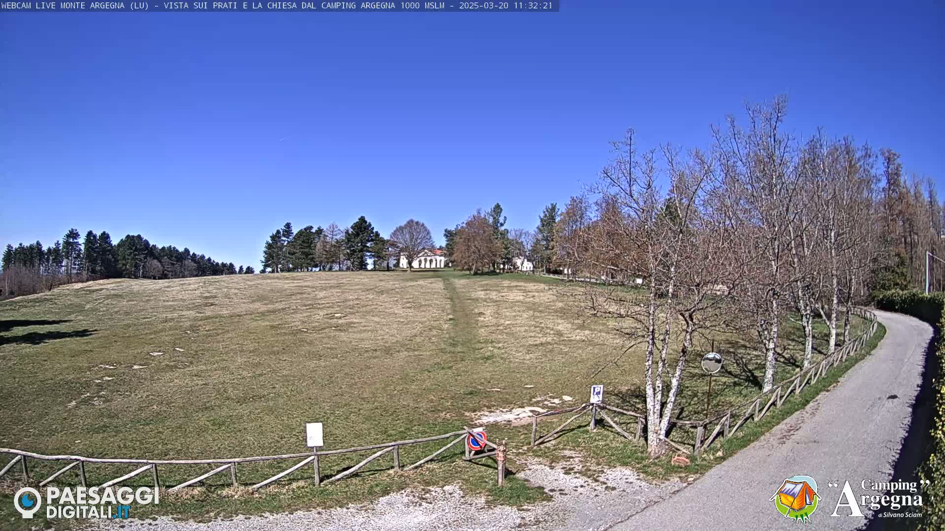A grassy field with a building in the distance, a road, and bare trees under a clear blue sky.