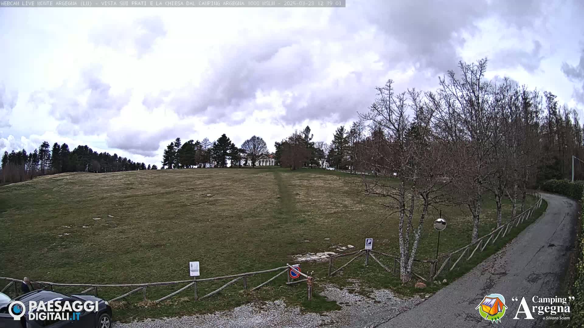 A mostly cloudy day reveals a grassy field leading to a building atop a hill, with leafless trees and a paved road curving through the foreground.