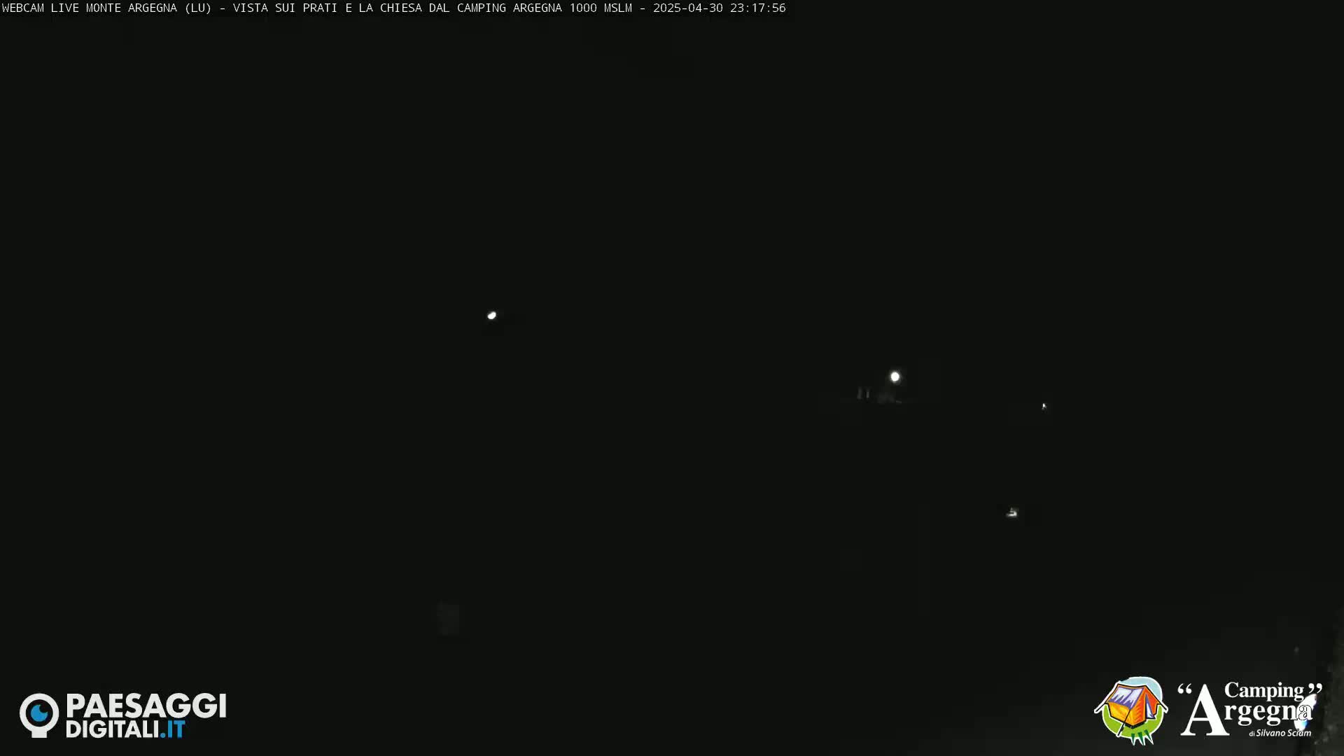 A nighttime, dark scene shows a few faint lights in the distance under a clear sky.