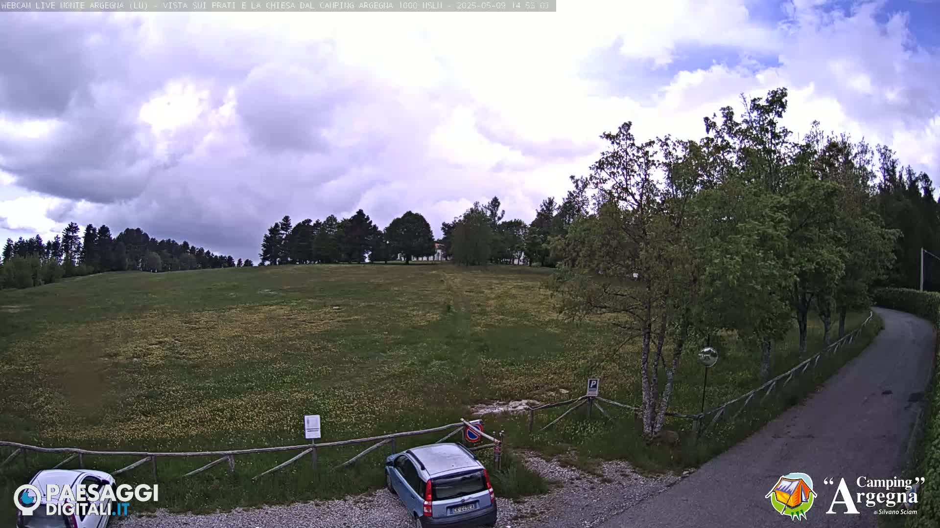 A mostly cloudy sky overlooks a grassy field with trees and a car parked in a small lot next to a curving road.