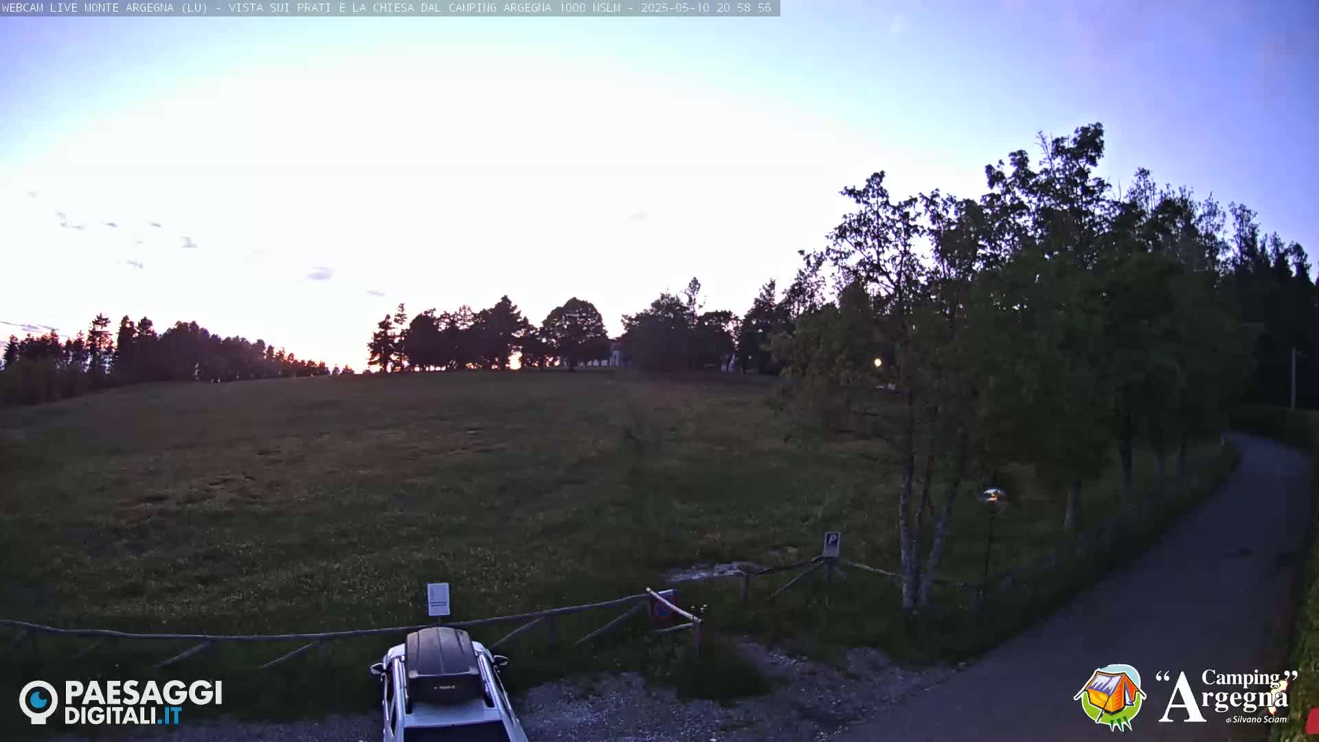 A twilight view shows a grassy field bordered by trees, a car in a parking area, and a paved road curving away from the camera.