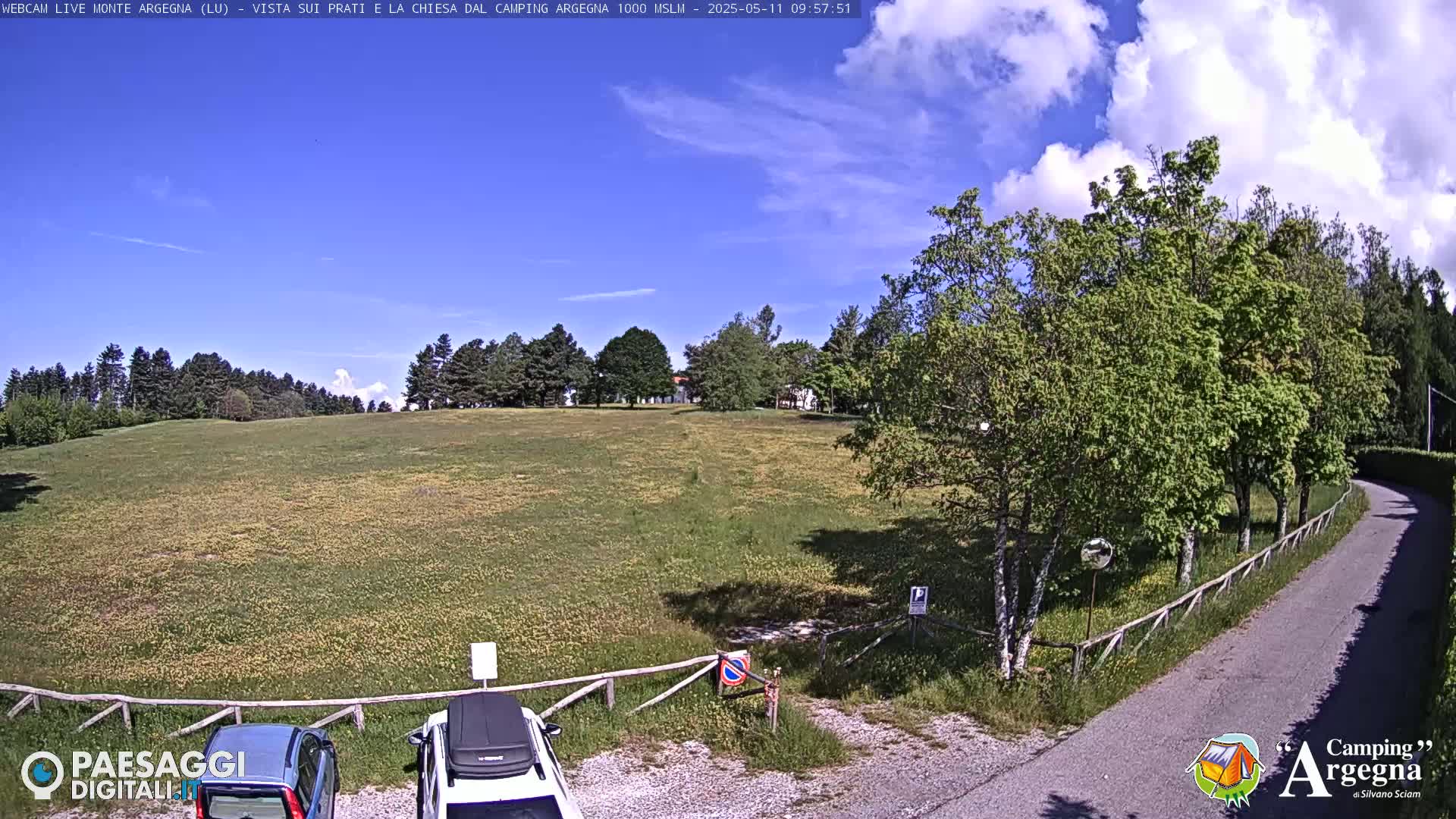 A mostly sunny day reveals a grassy field bordered by trees and a paved road curving into the scene, with two cars parked near a wooden fence.