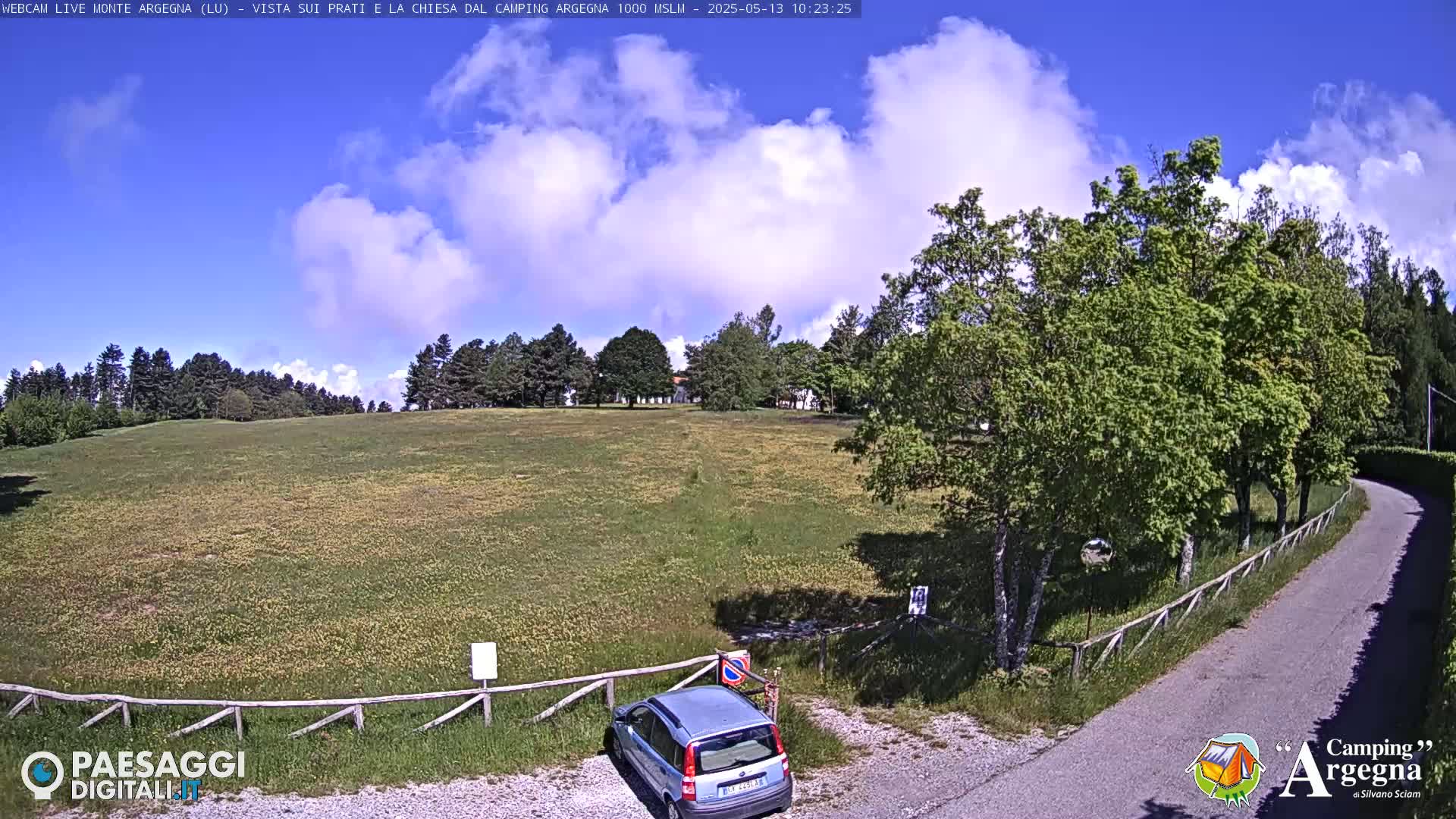 A partly cloudy, sunny day shows a grassy field with a few trees and a small car parked near a curving road.