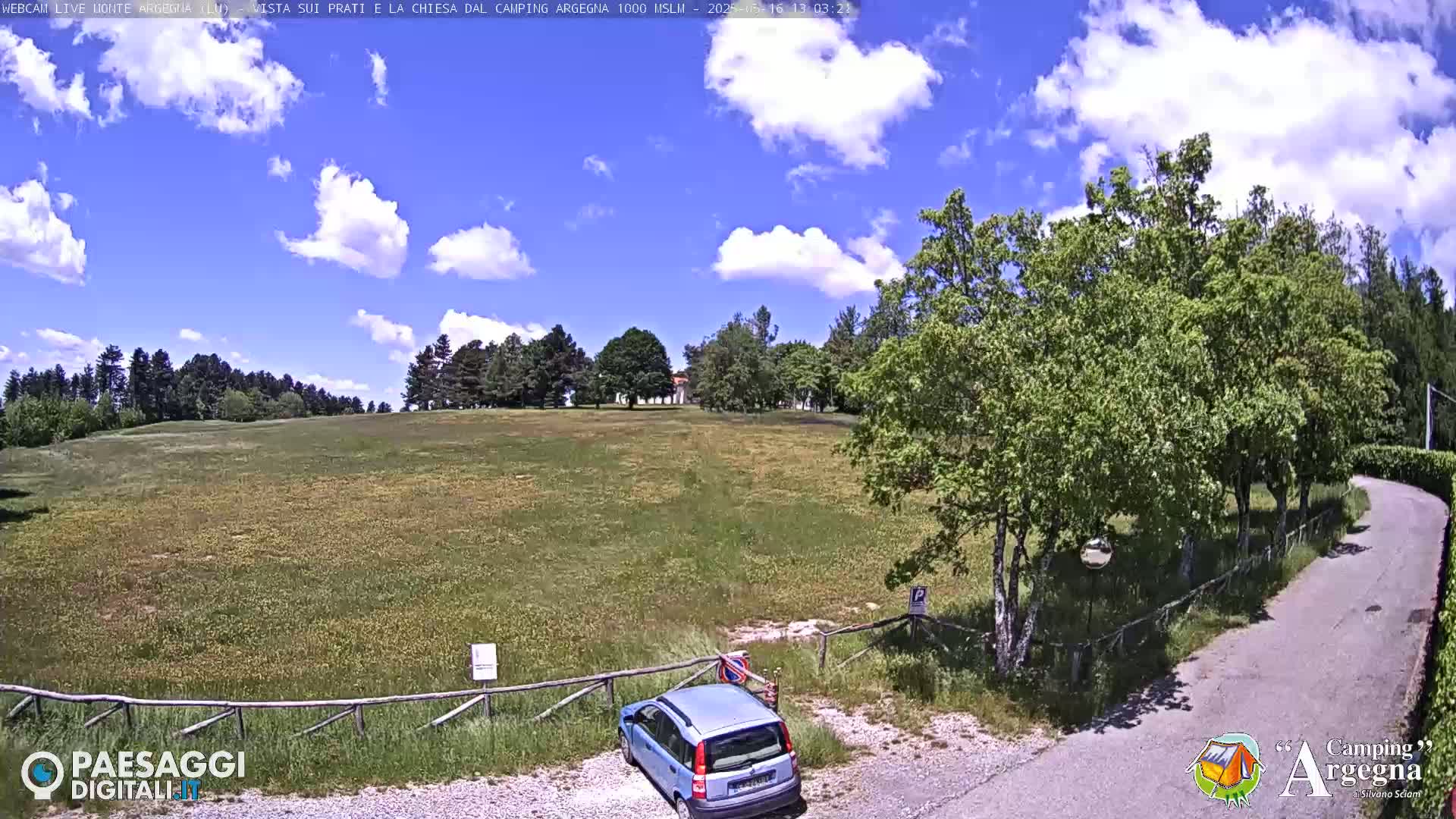 A light blue station wagon is parked near a road that leads to a grassy field under a partly cloudy blue sky.