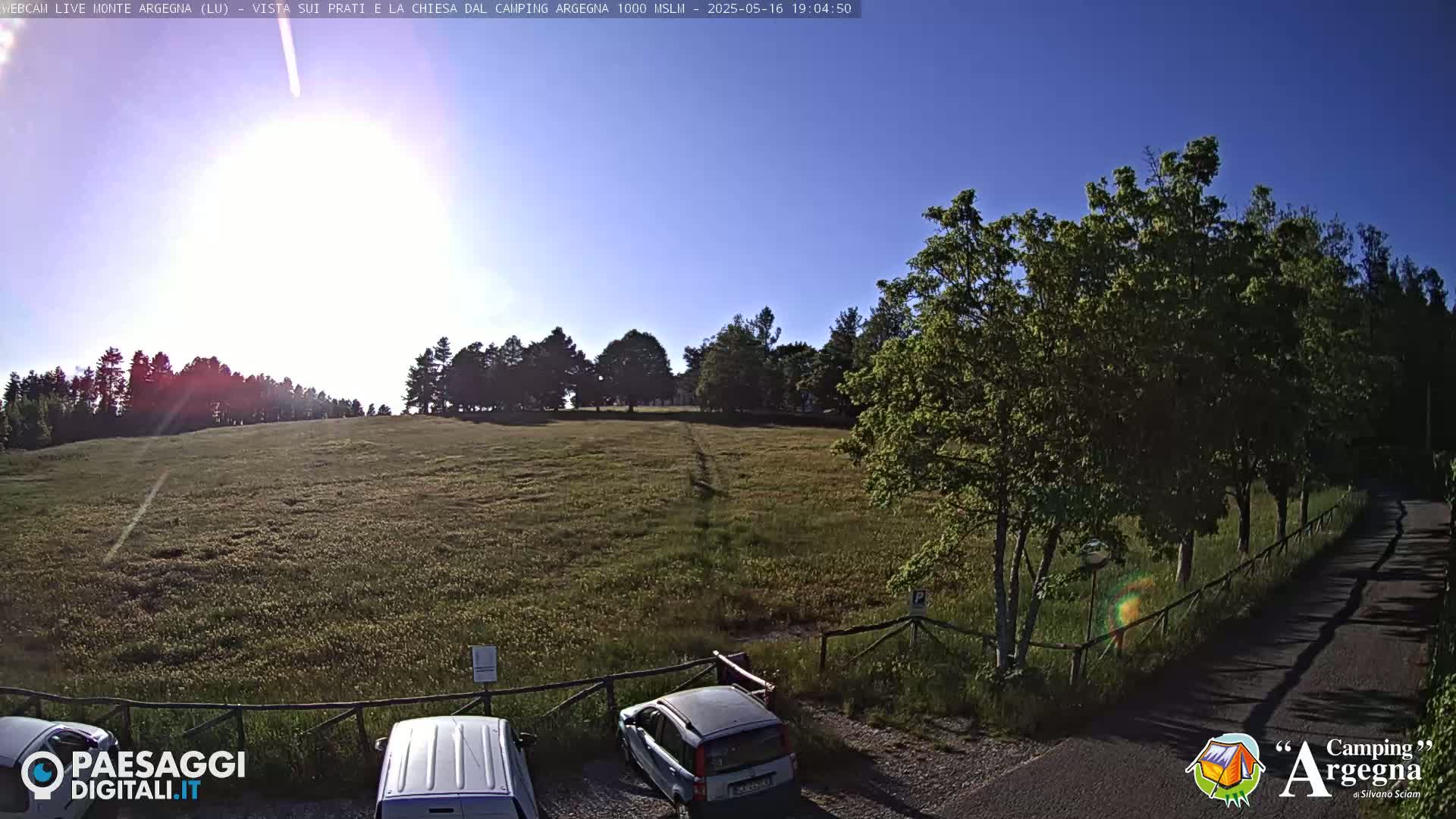 A sunny day reveals a grassy field bordered by trees, a paved road, and a small parking area with two cars.