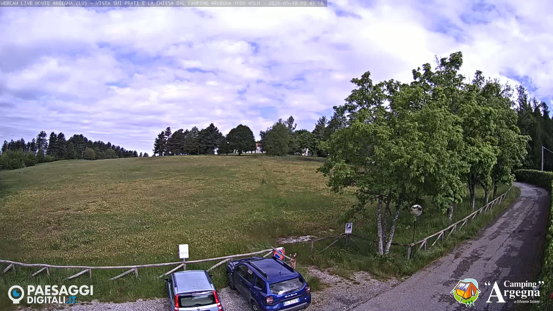 A partly cloudy day reveals a grassy field, bordered by trees and a wooden fence, with two cars parked in a small lot near a roadway.
