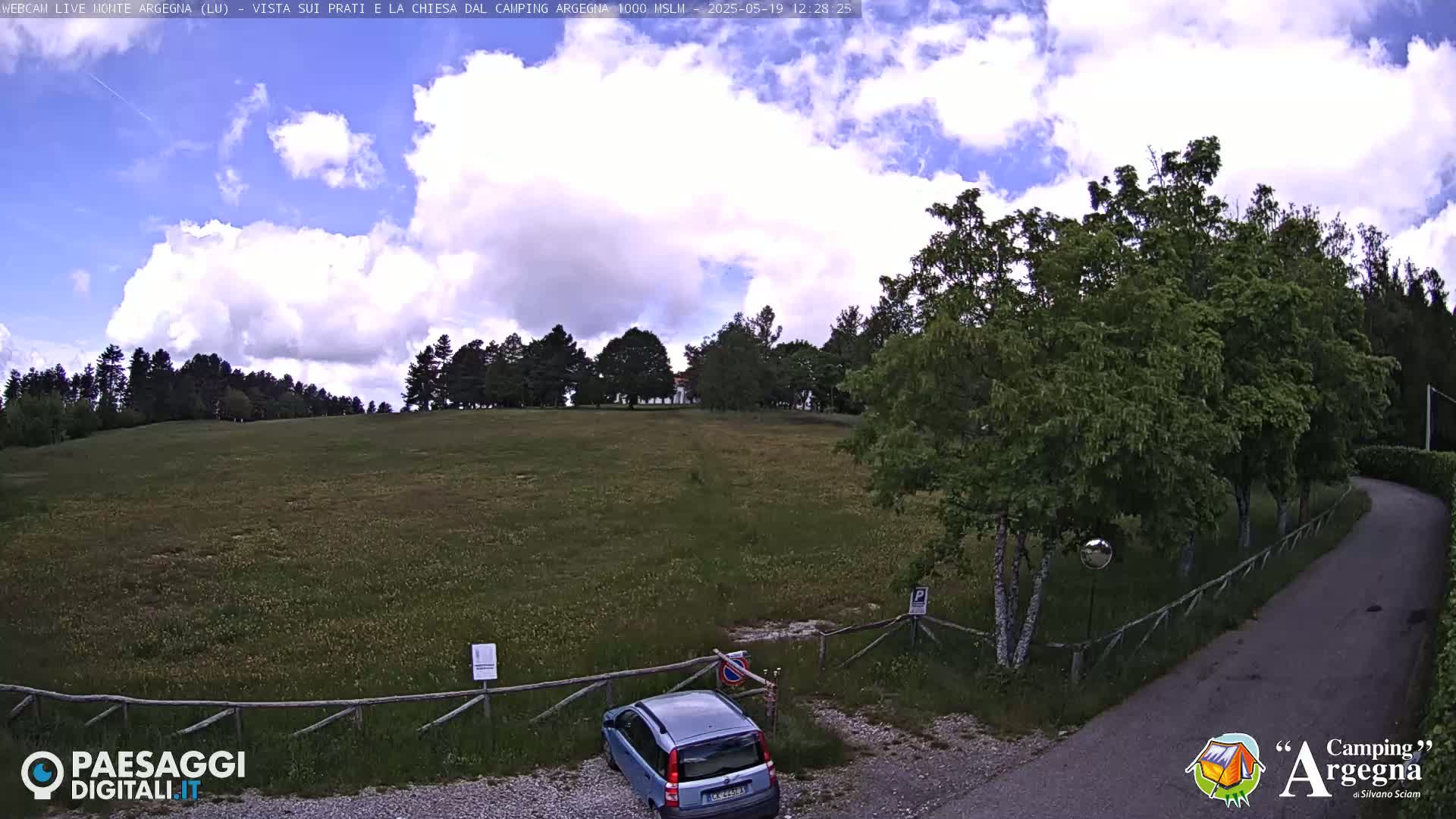 A partly cloudy sky overlooks a grassy field with trees in the background, a small car parked near a wooden fence, and a paved road leading to the right.
