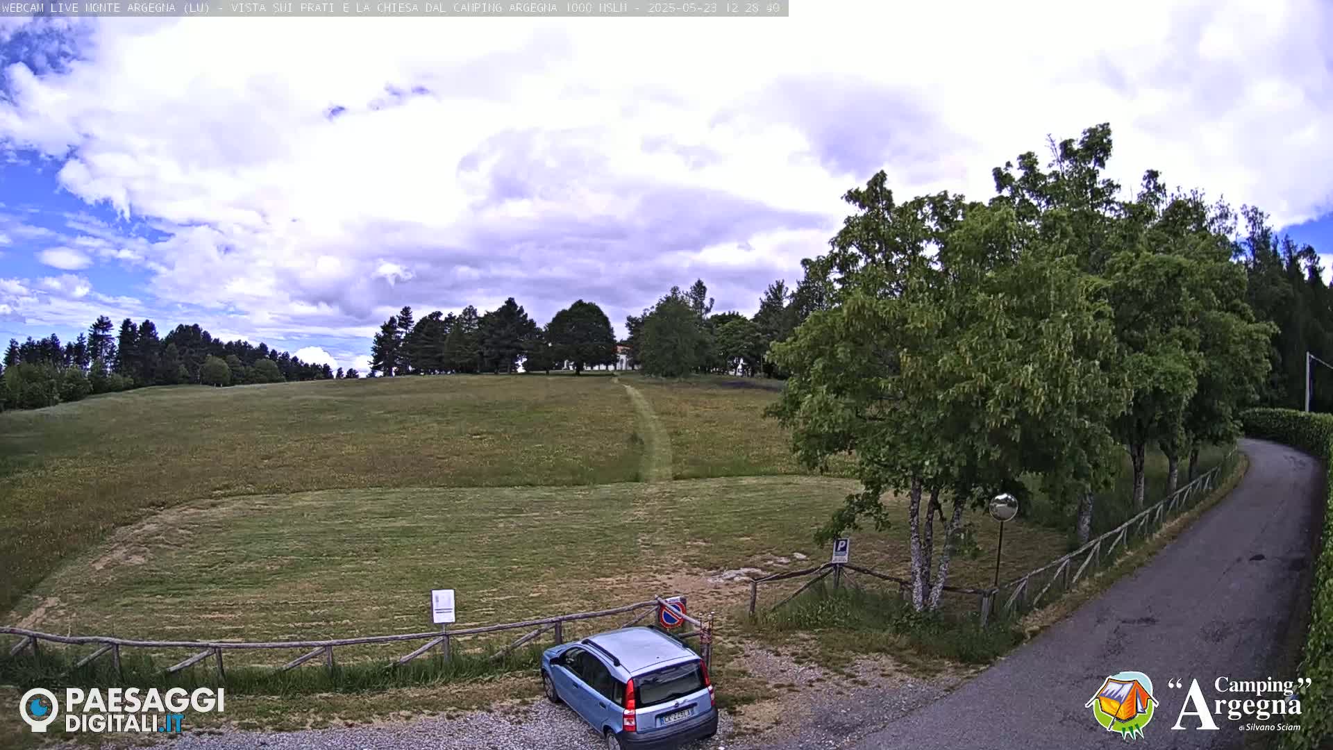 A partly cloudy sky overlooks a grassy field with a small blue car parked near a curving road, bordered by trees and a wooden fence.