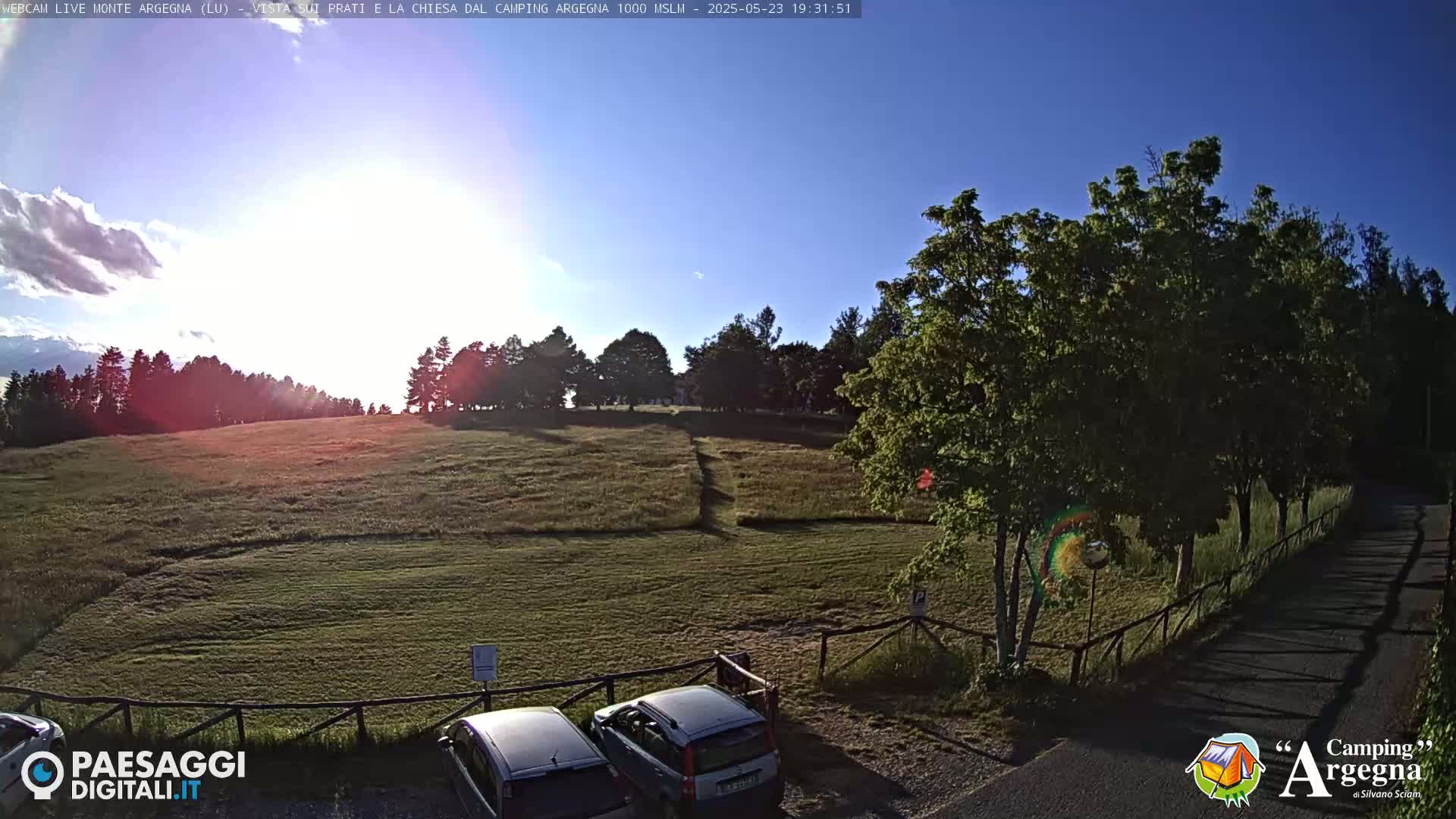 A grassy field with a few parked cars is bordered by trees under a mostly sunny sky with some scattered clouds.