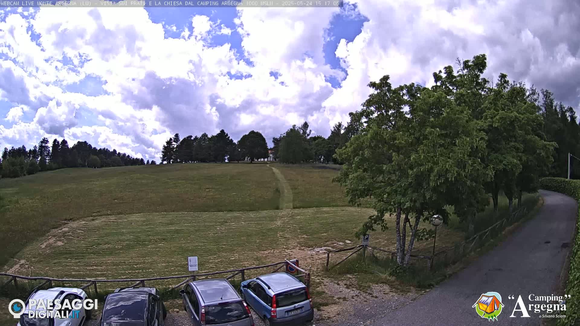 A partly cloudy sky overlooks a grassy field with a few cars parked near some trees and a road curving to the right.