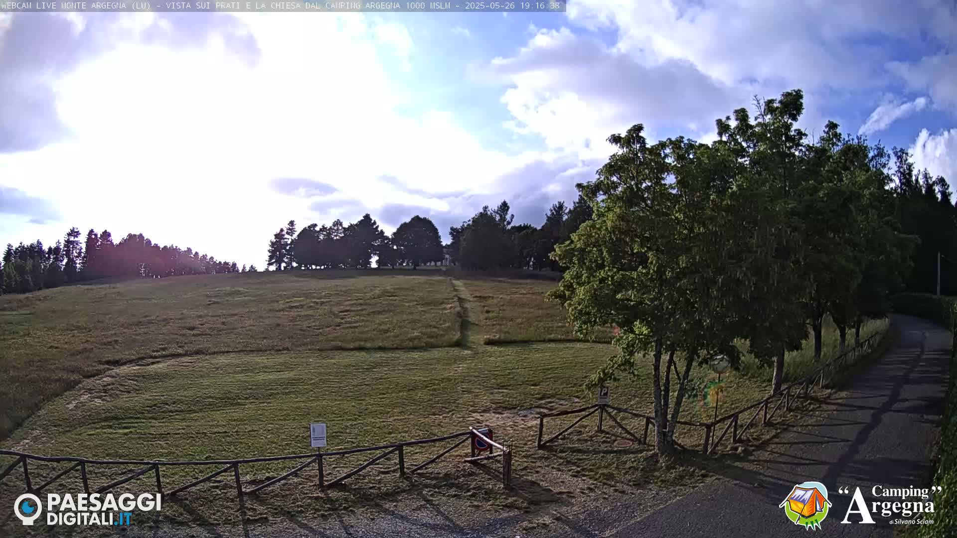 A grassy field with a wooden fence and a paved road, partially shaded by trees, under a partly cloudy sky.