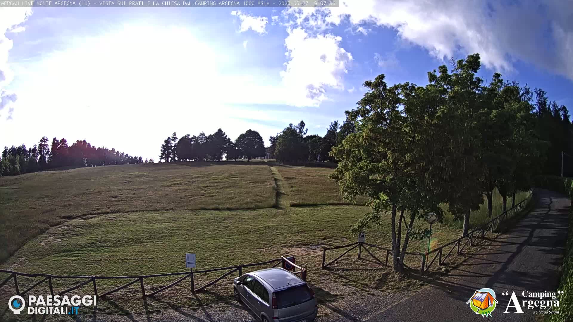 A mostly sunny day with partly cloudy skies shows a grassy field with a few trees, a car parked near a wooden fence, and a paved road leading away.