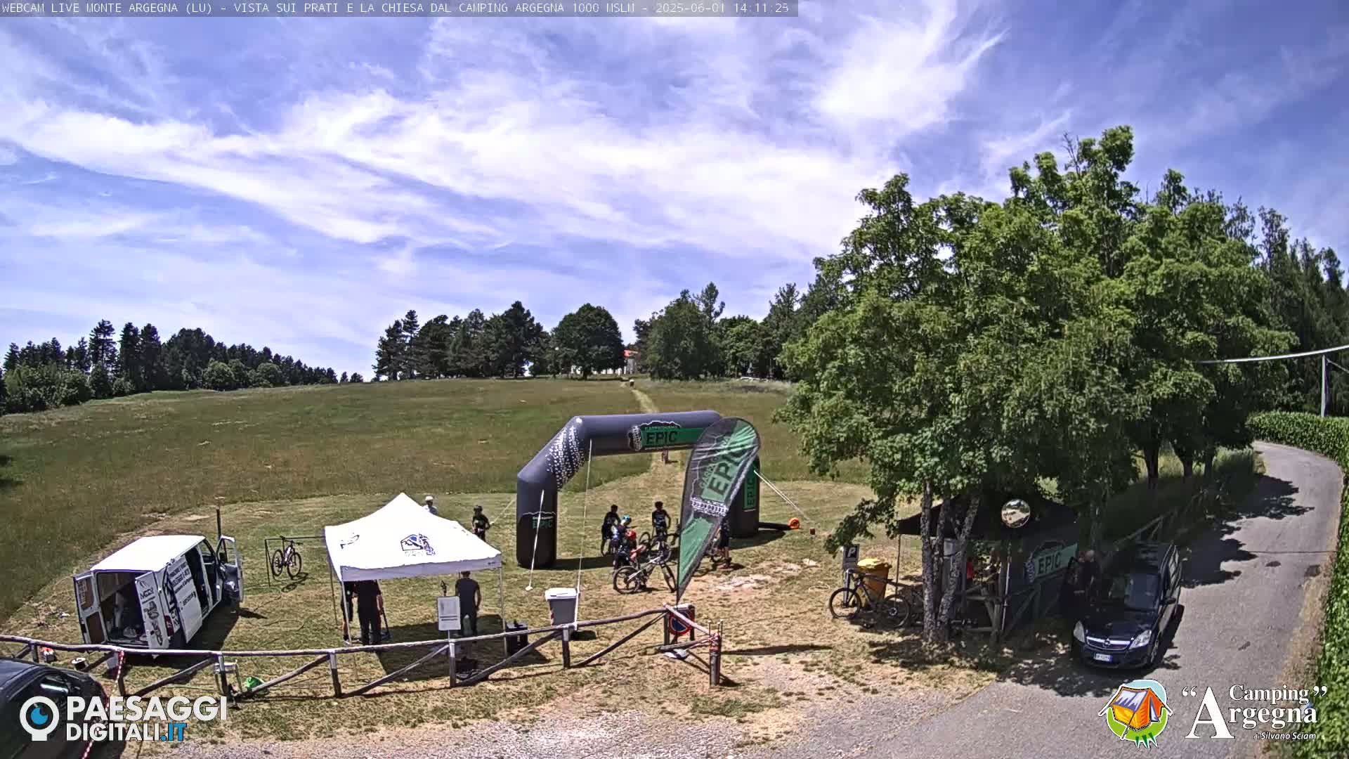 A sunny outdoor scene shows a mountain bike race finish line with several people, tents, vehicles, and bicycles in a grassy field bordered by trees.