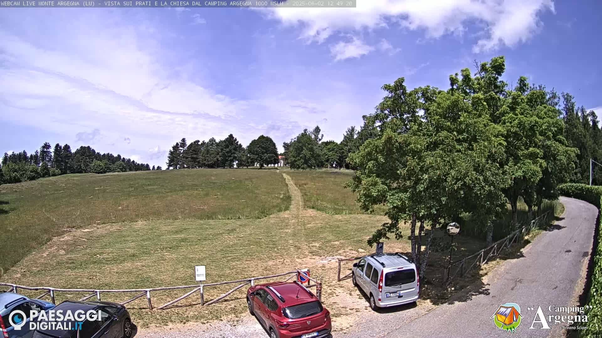 A grassy field with a dirt path leading to a small building is partially visible, with cars parked near a wooden fence under a partly cloudy sky.