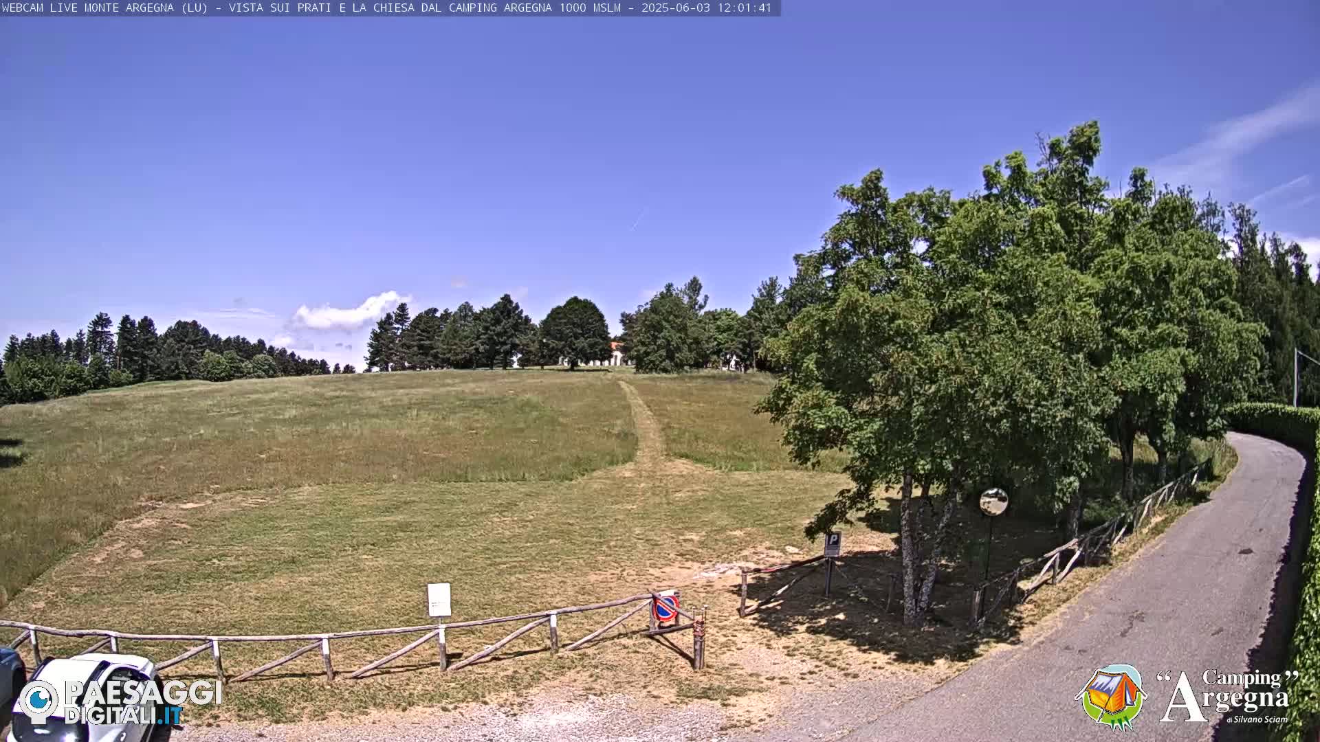 A grassy field with a path leading to a building in the distance, under a mostly clear blue sky, is surrounded by trees and a paved road.