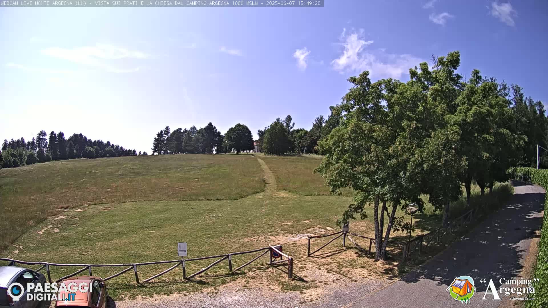 A grassy field with a dirt path leading to a small building is visible under a mostly clear, sunny sky; trees line the horizon and border the field, and a wooden fence is in the foreground.