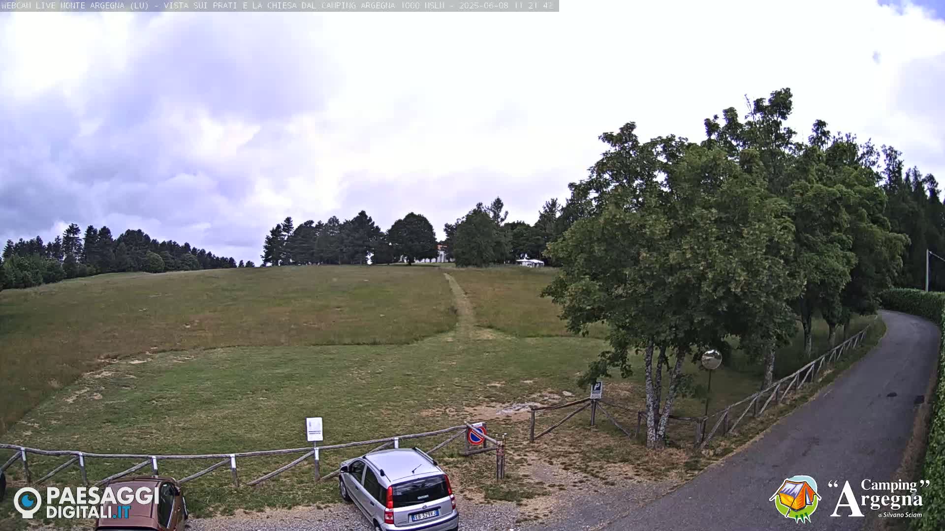 A mostly cloudy day reveals a grassy field with a dirt path leading to a line of trees in the distance, a car parked in a lot, and a road curving to the right.