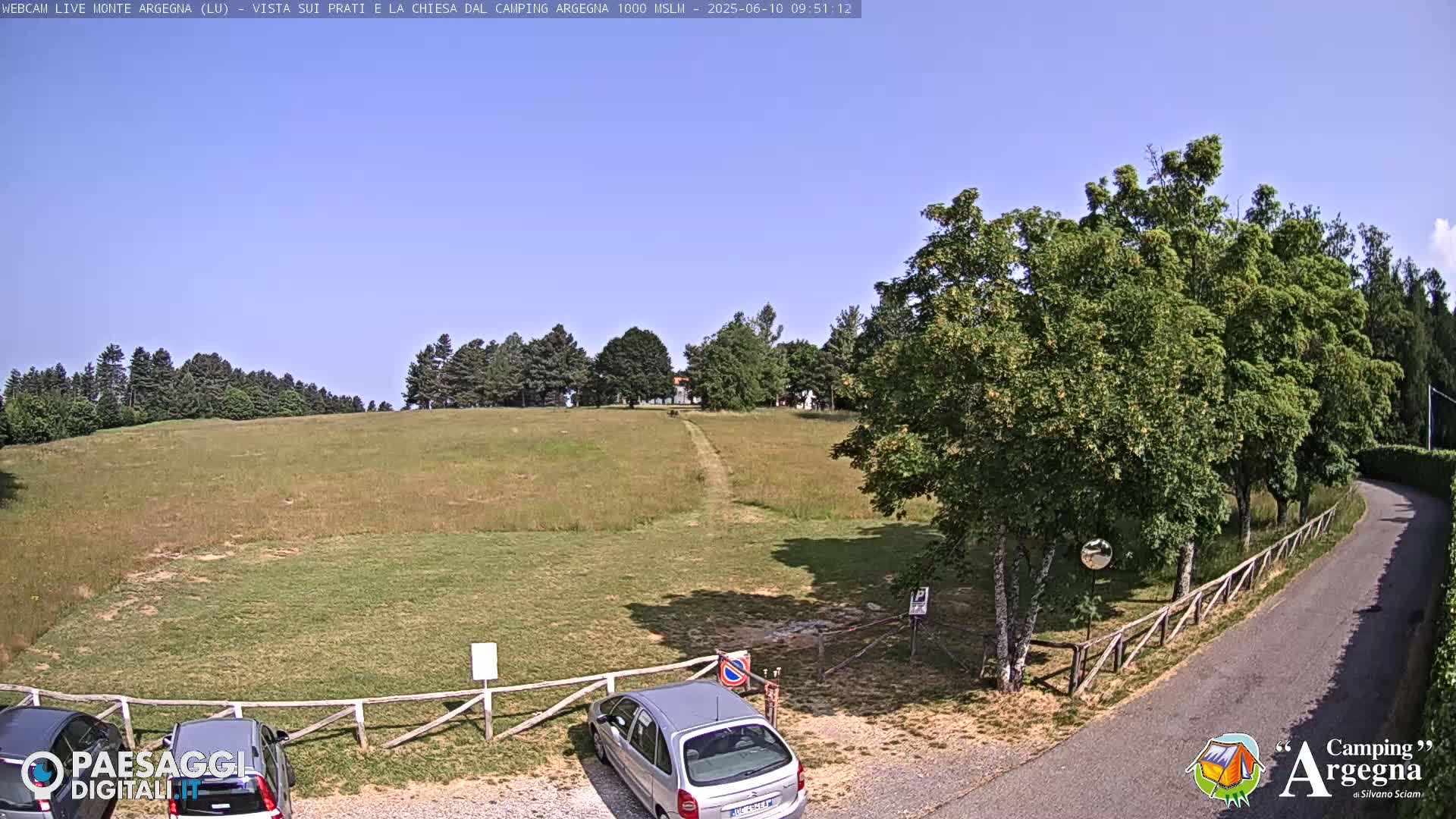 A grassy field with a dirt path leading to a line of trees under a clear blue sky, a few cars parked near a wooden fence beside a paved road.