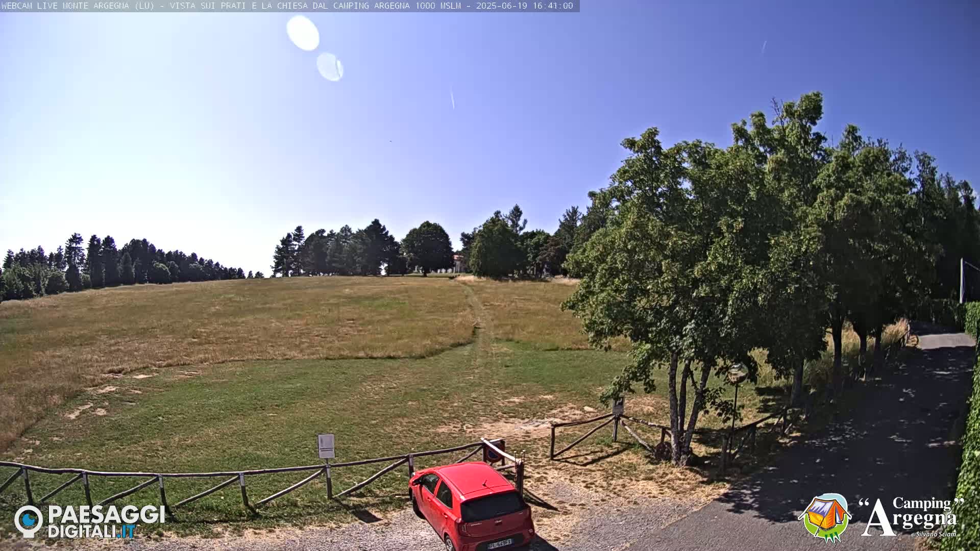 A red car is parked near a wooden fence in a grassy field, under a clear blue sky.