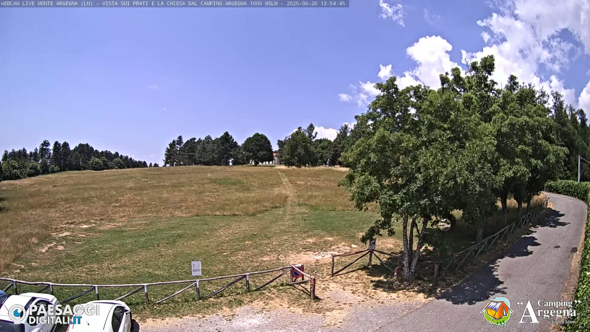 A grassy field with a dirt path leading to a small building, surrounded by trees under a partly cloudy blue sky.