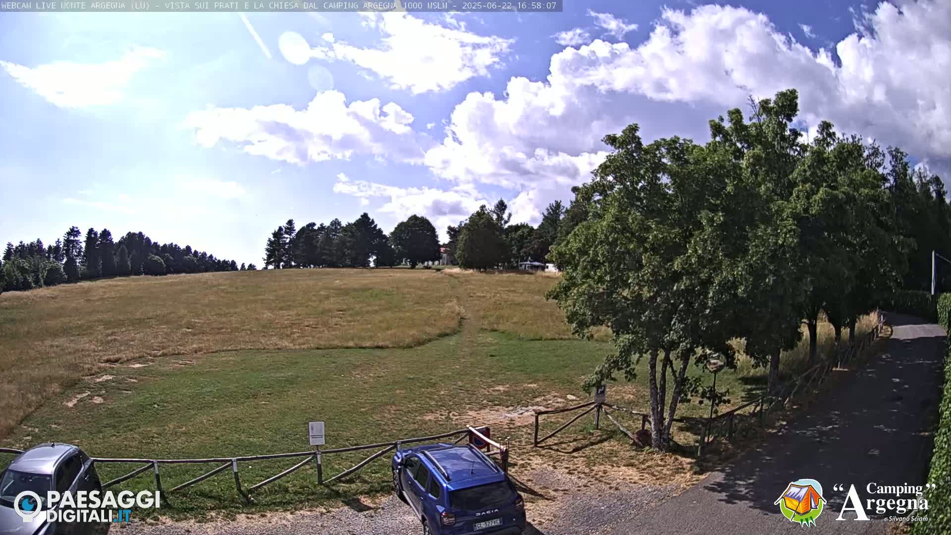 A partly cloudy sky overlooks a grassy field with a few trees and parked cars near a gated entrance.