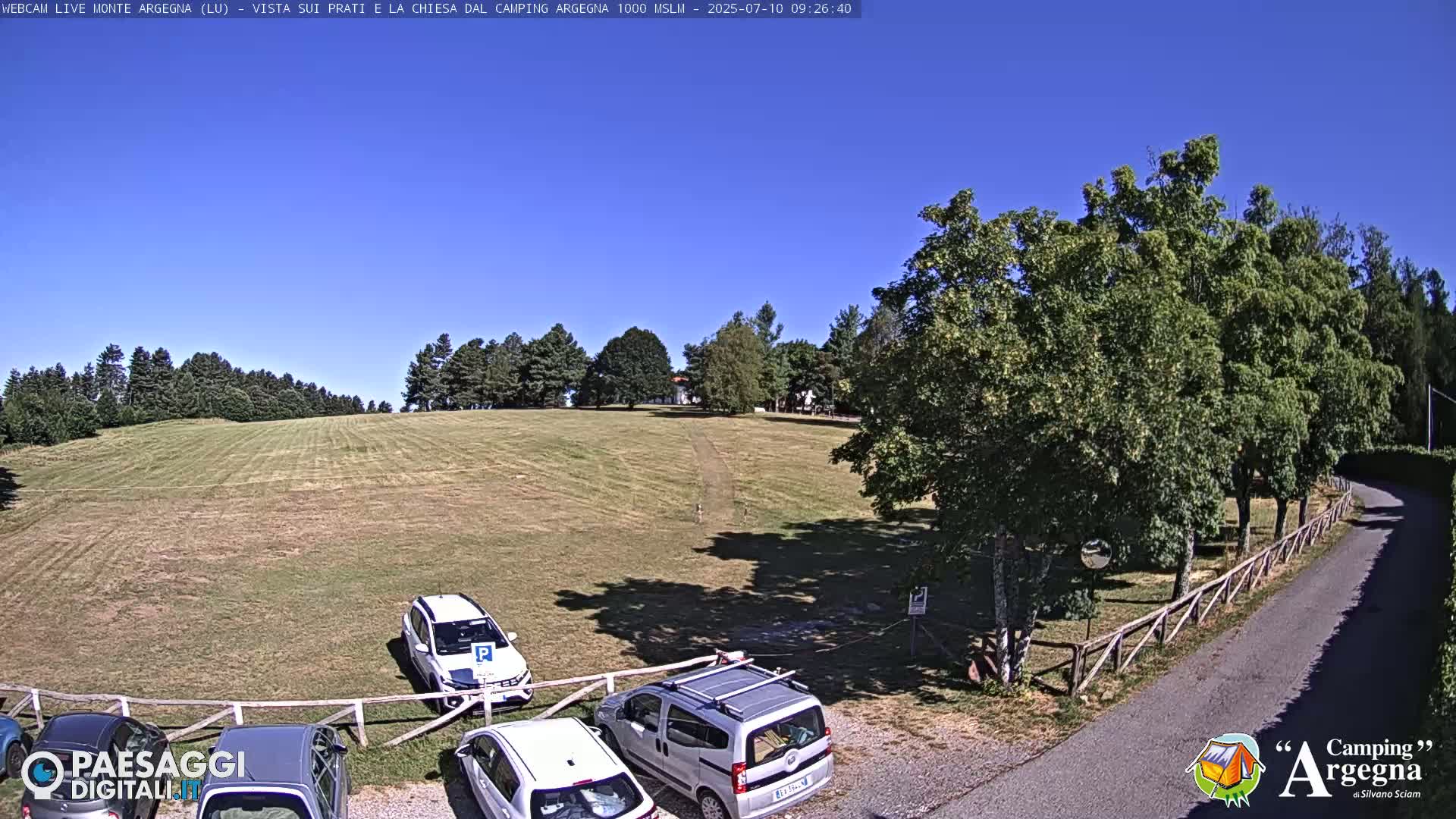 A grassy field with several parked cars is visible beneath a clear blue sky, bordered by trees and a paved road.