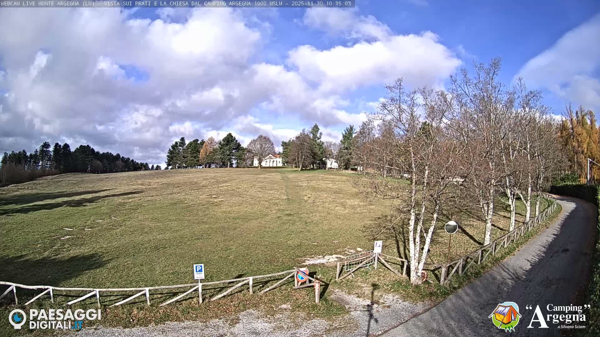 A wide, gently sloping grassy field stretches towards distant trees and a few white buildings under a partly cloudy sky, with a paved road and bare-branched trees lining the right foreground on a bright day.
