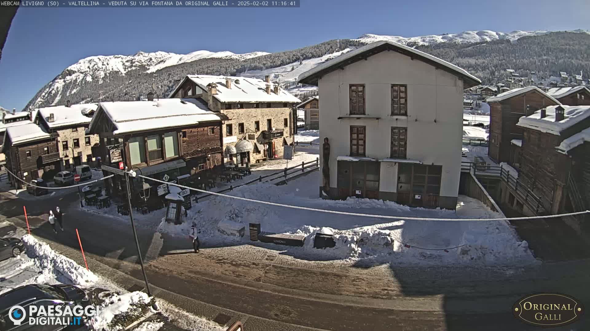 A snow-covered village square, nestled amongst snow-dusted buildings and mountains under a sunny sky.