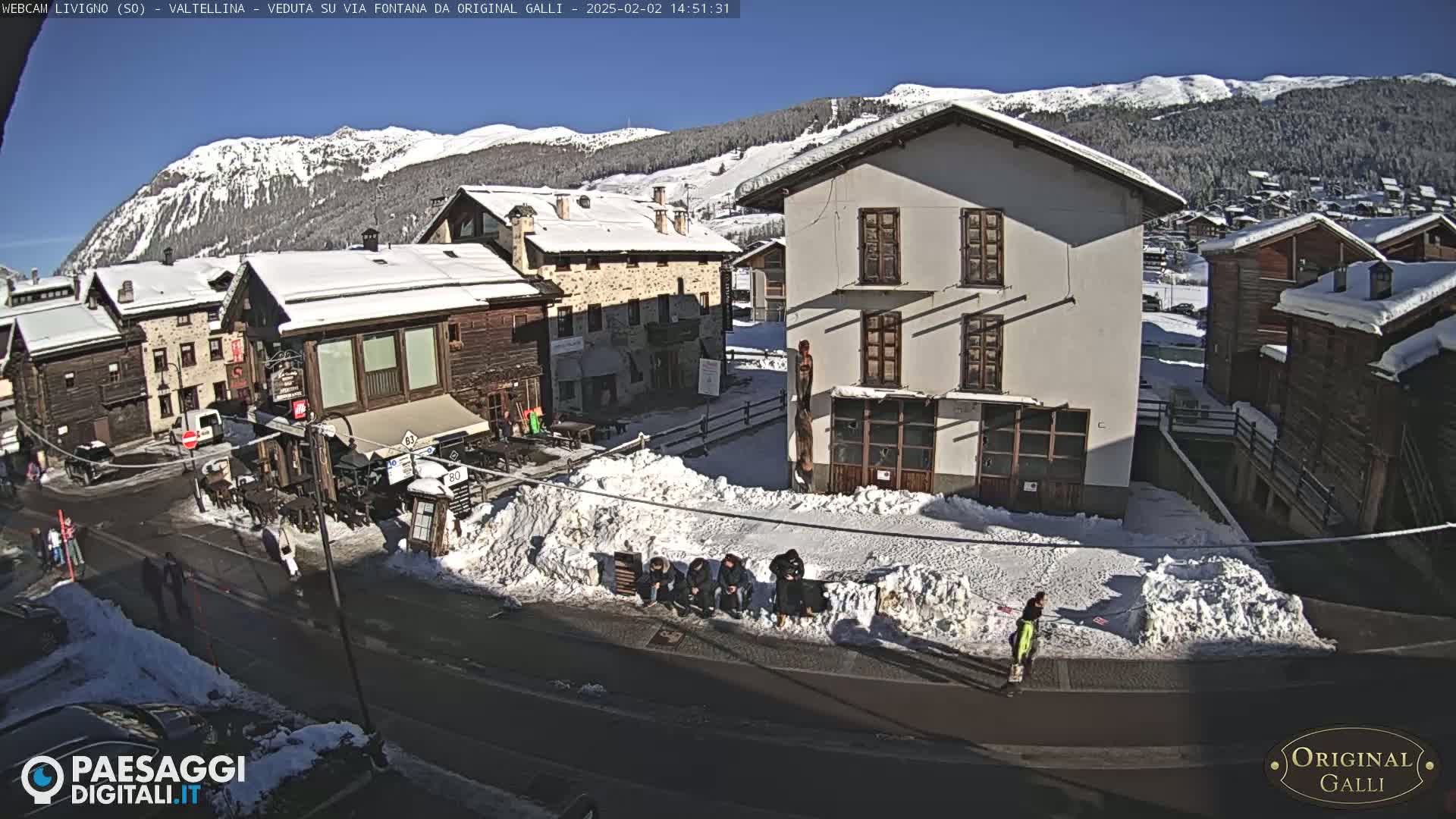A sunny day in a snow-covered village shows several buildings, a street, and a few people standing near snow piles.