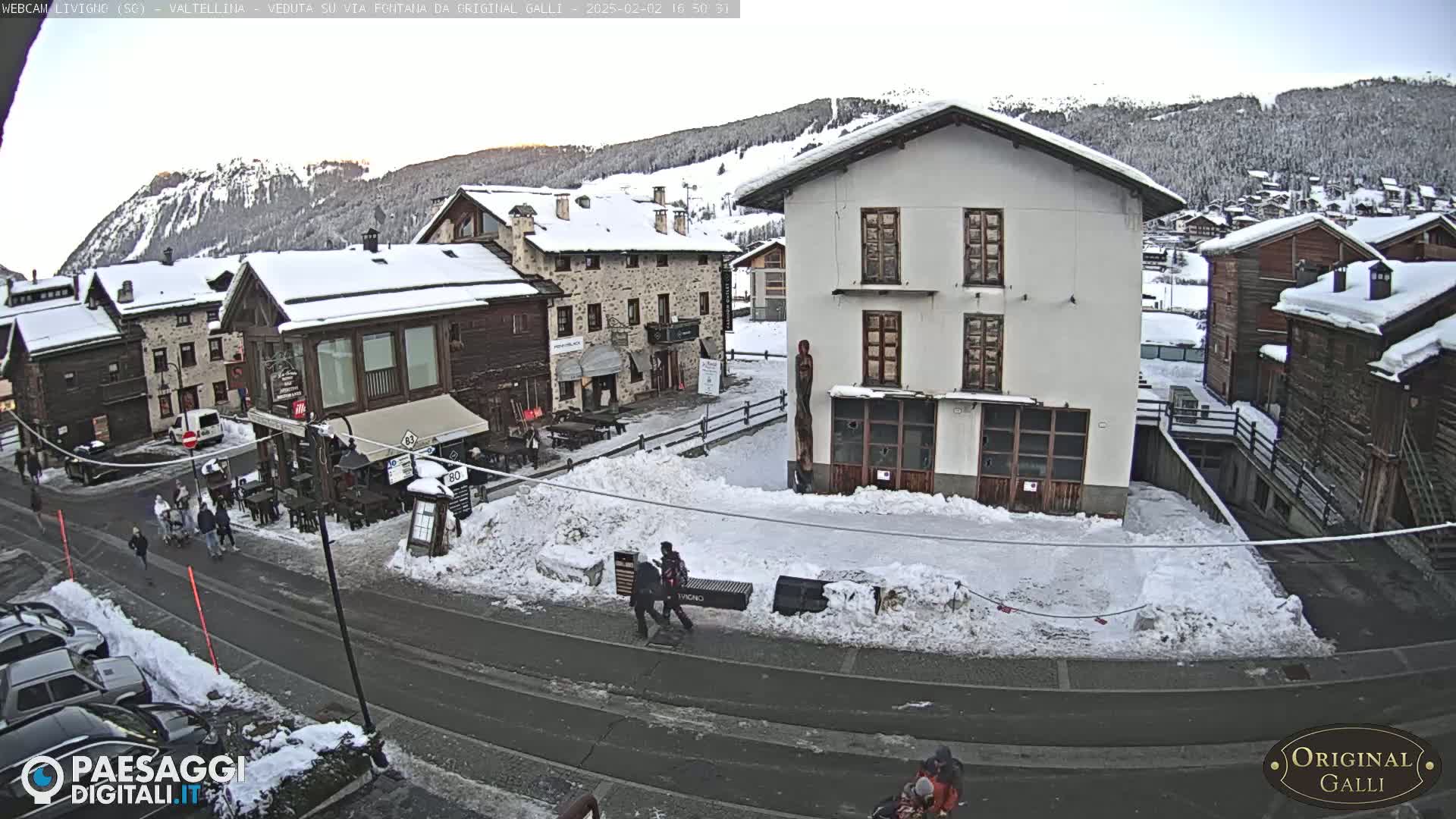 A snow-covered village street scene with several buildings, some pedestrians, and parked cars, under a mostly clear sky.
