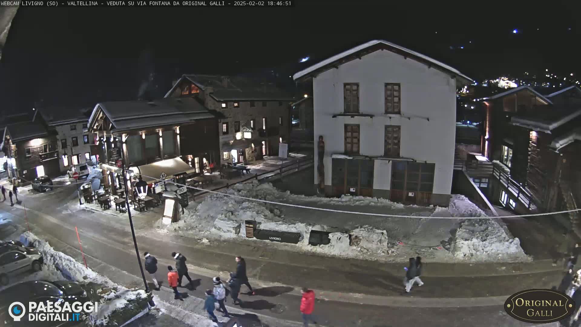 A snow-covered town square at night, with several people walking along a street lined with buildings.