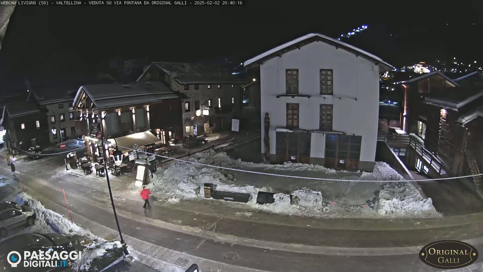 A snow-covered street scene at night shows several buildings, one of which is a restaurant with outdoor seating, and a few people walking.