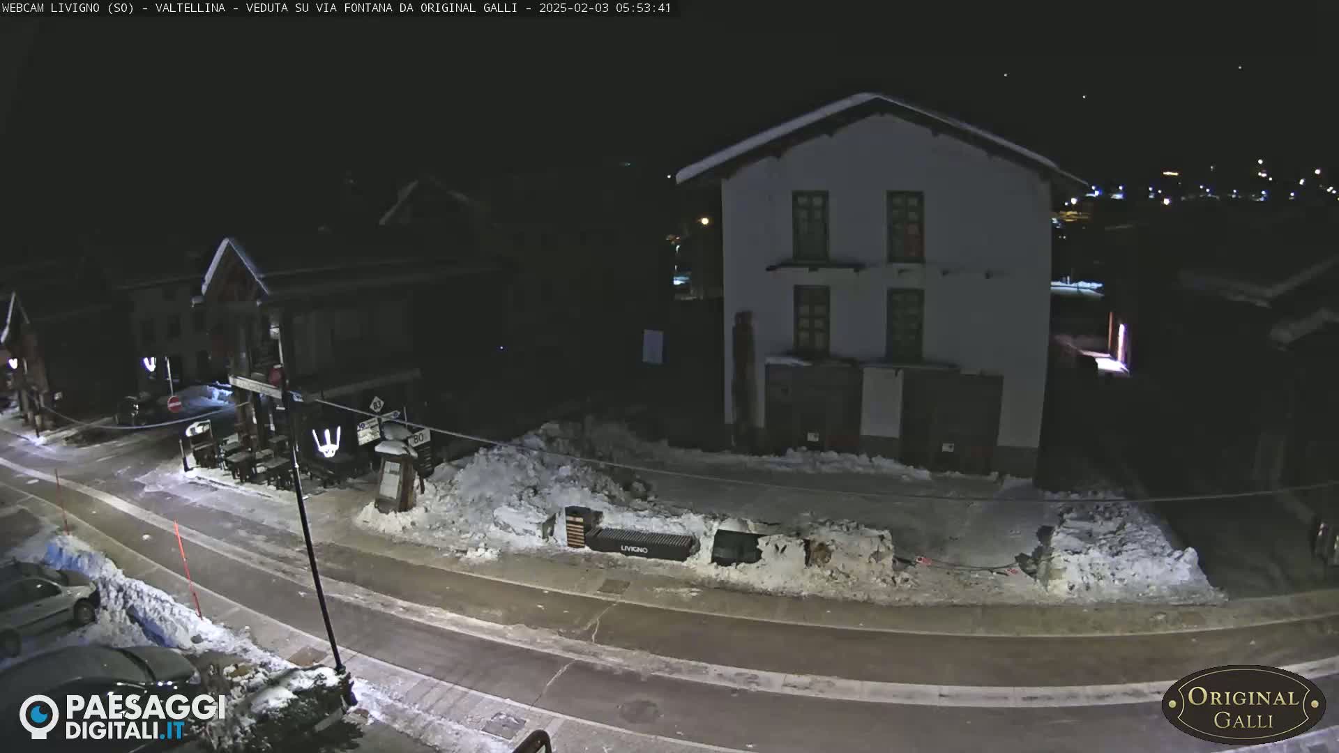A nighttime street scene in a snowy village shows a two-story building and a road with parked cars.