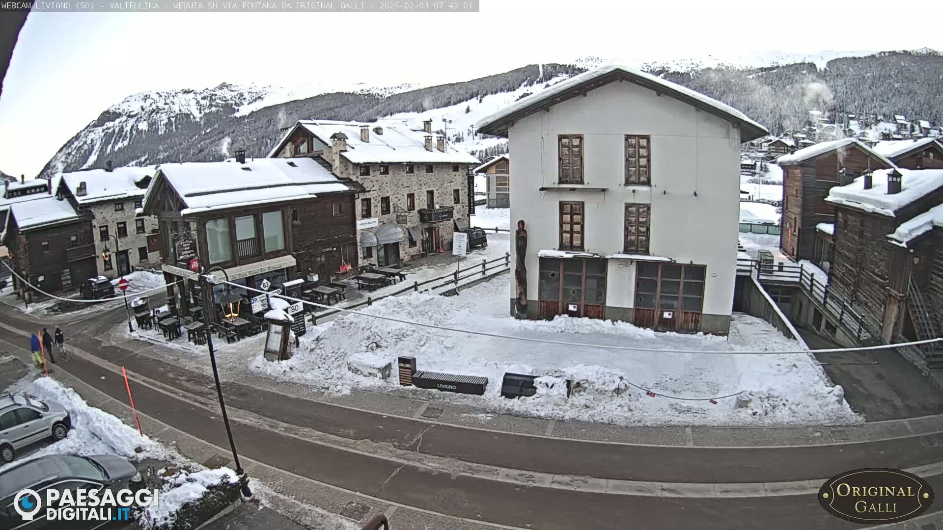 A snow-covered village street scene with several buildings, some with outdoor seating areas, under an overcast sky.