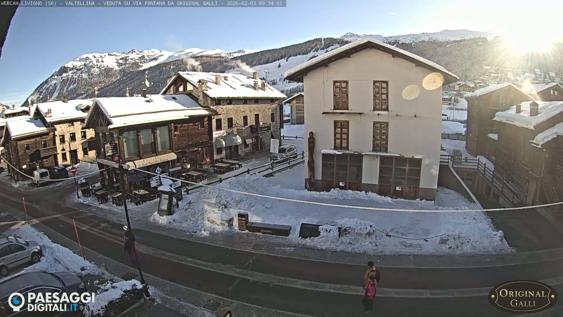 A sunny, snow-covered village square with several buildings, some cars, and a few pedestrians, set against a backdrop of snow-capped mountains.