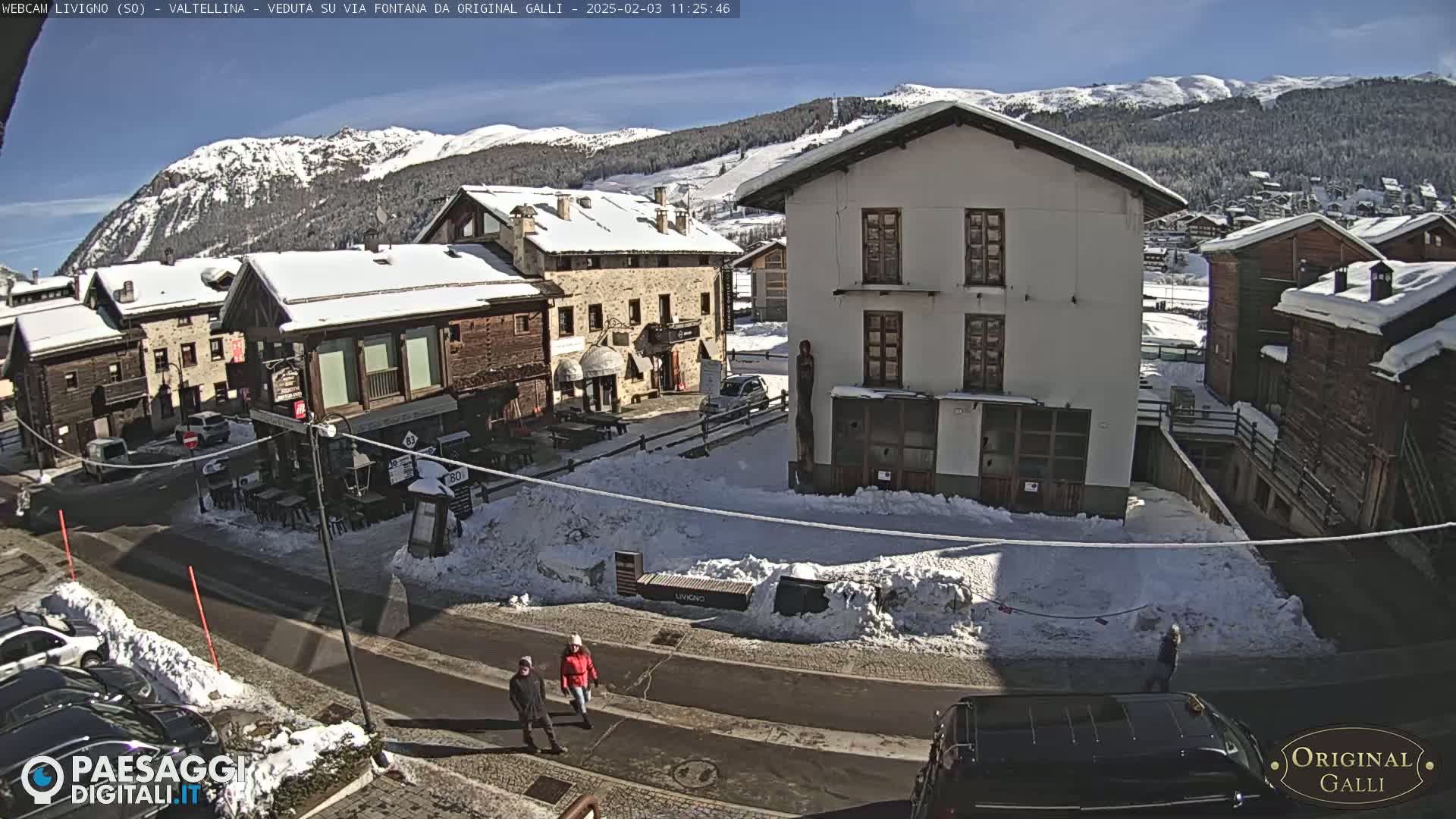A snow-covered village square with several buildings, a few pedestrians, and cars parked along the street under a sunny sky.