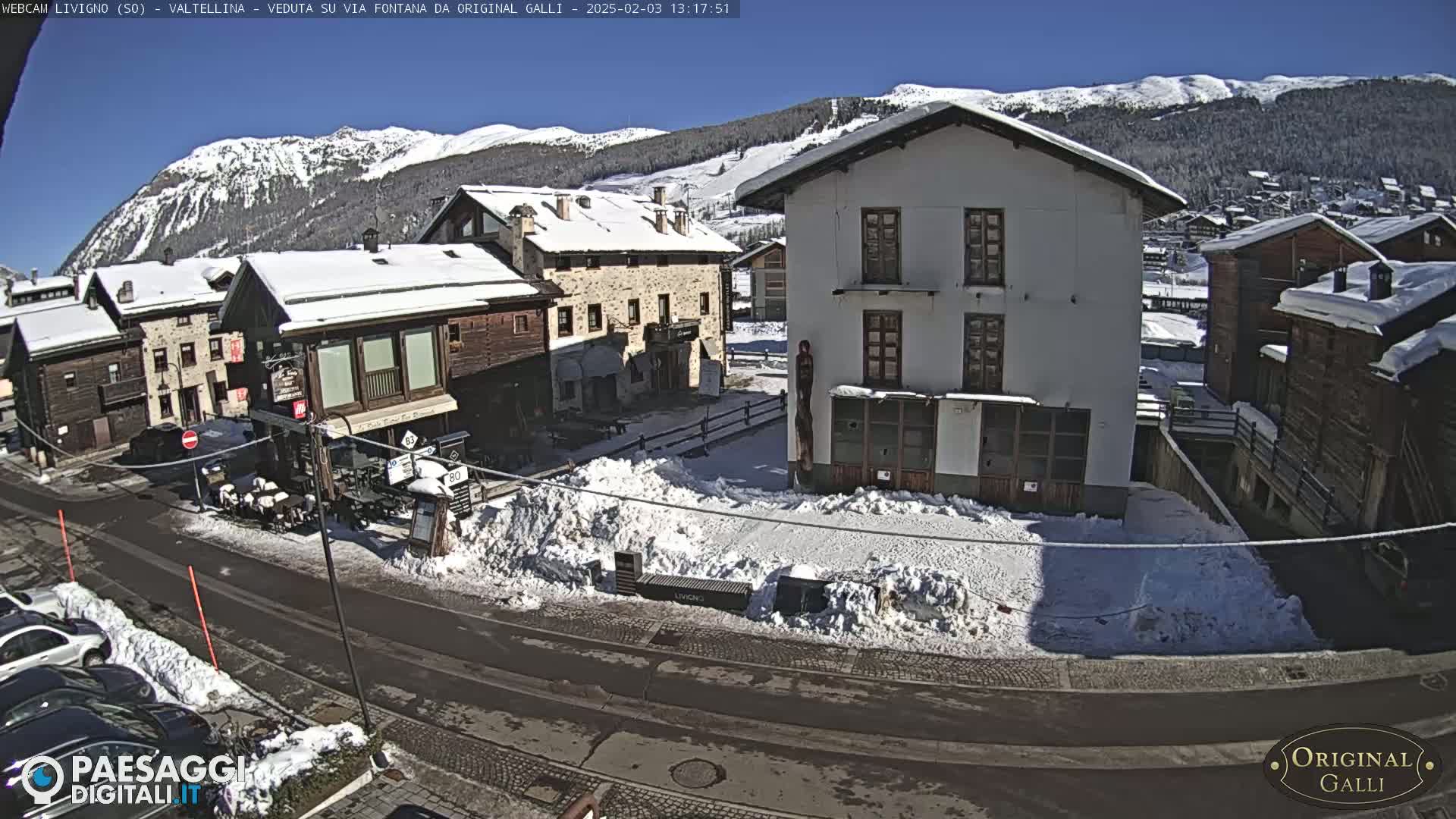 A snow-covered village scene on a sunny day, showing several buildings and a street with parked cars.