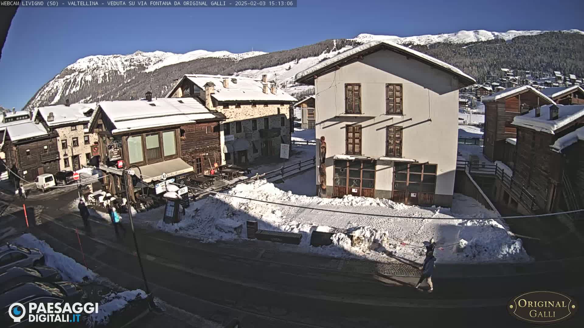 A snow-covered village street scene on a sunny day, with several buildings, some cars, and a few pedestrians.