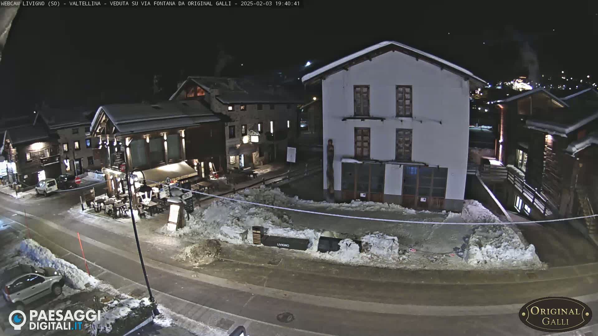 A snow-covered town square at night, with several buildings illuminated, and cars parked along a road.