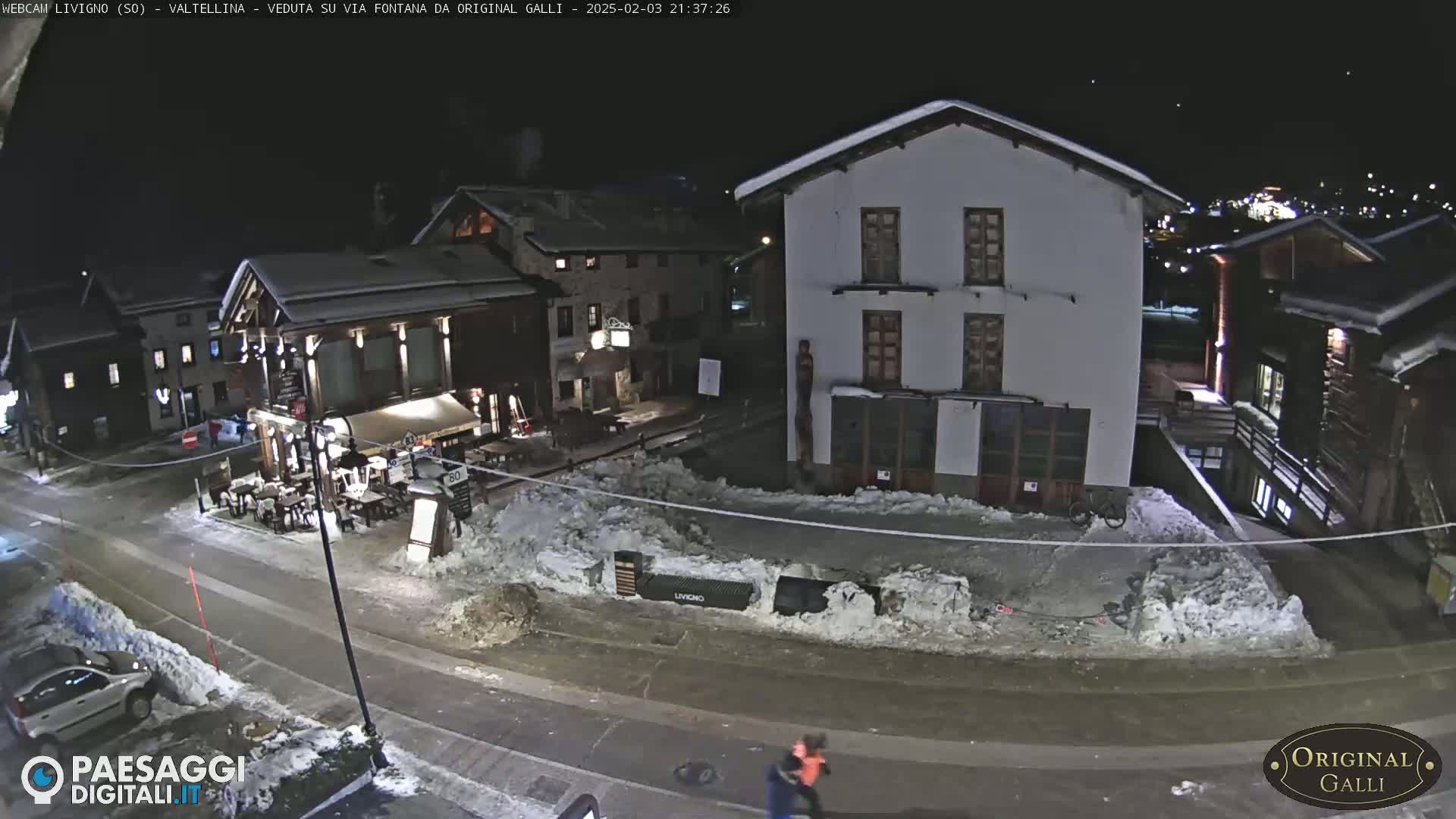 A nighttime view of a snow-covered village street with several buildings, one of which appears to be a restaurant with outdoor seating.