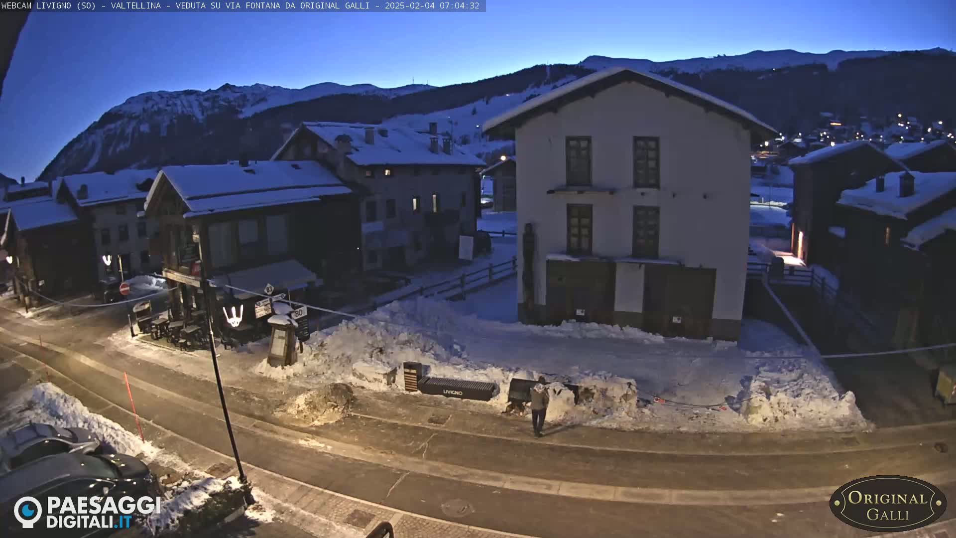 A snow-covered village street at twilight, with several buildings and a person walking near snowbanks, is nestled in a valley against a backdrop of snow-capped mountains under a dark blue sky.