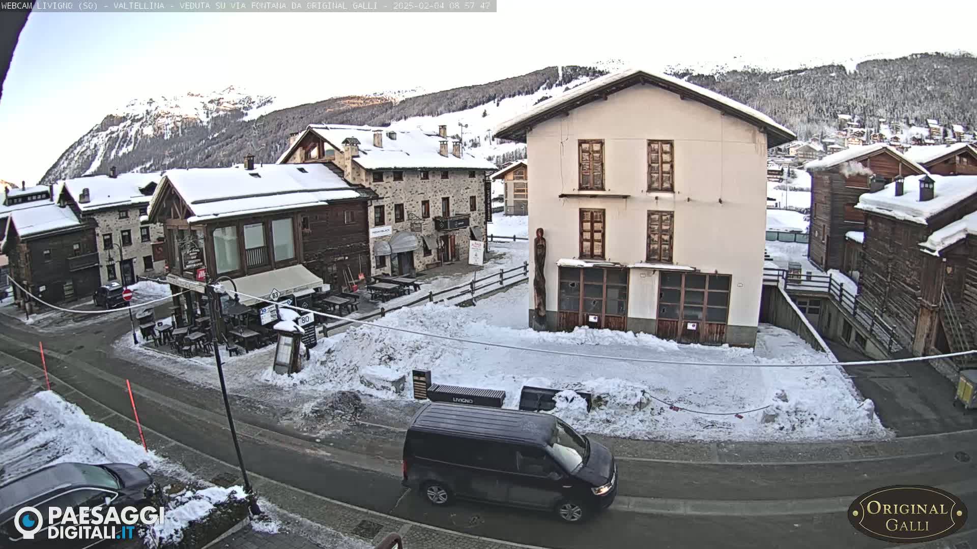 A snow-covered village street scene with several buildings, a dark-colored van, and a mountain range in the background under a mostly clear sky.